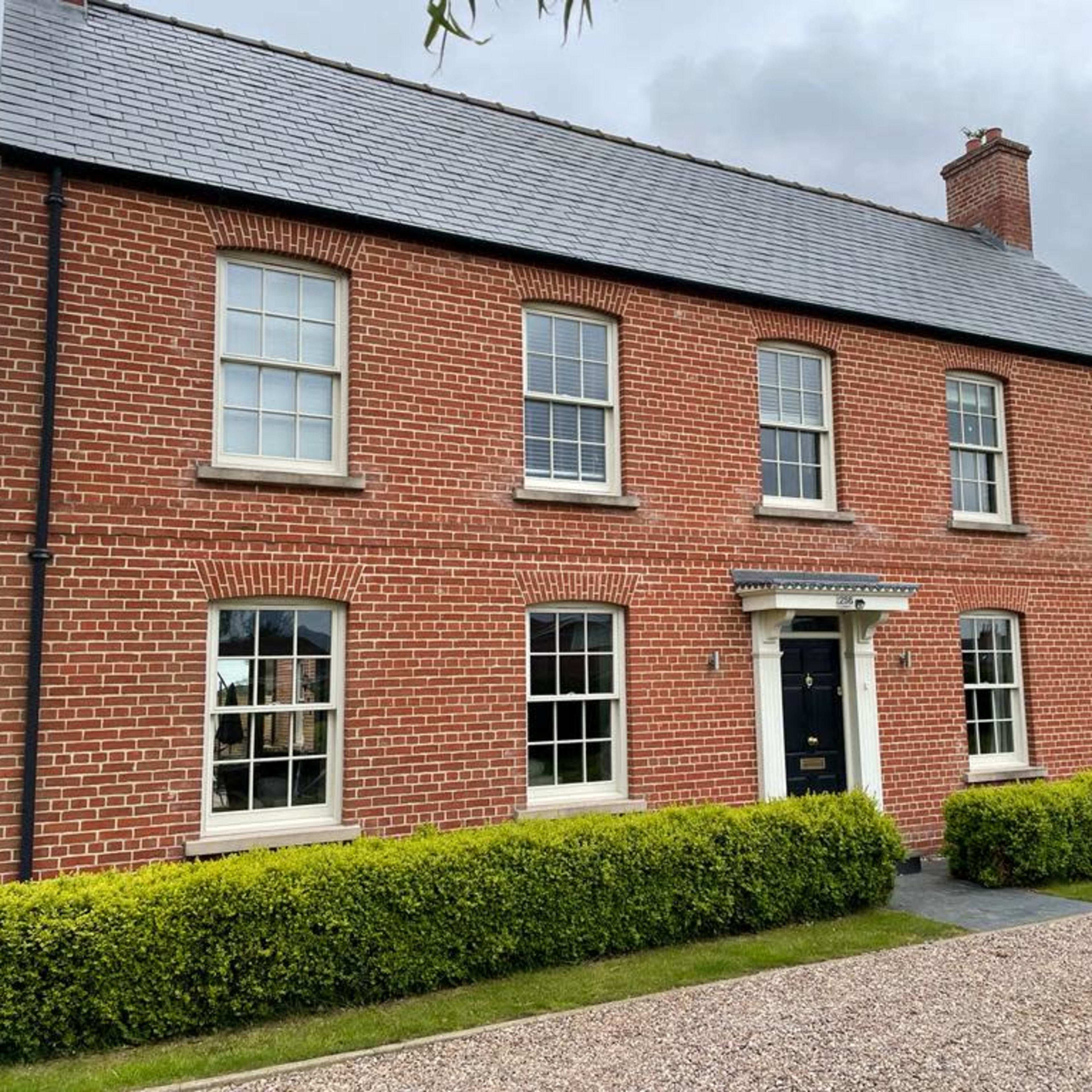 A red-brick house with white windows and a black front door is set against a gray sky and surrounded by neatly trimmed hedges.