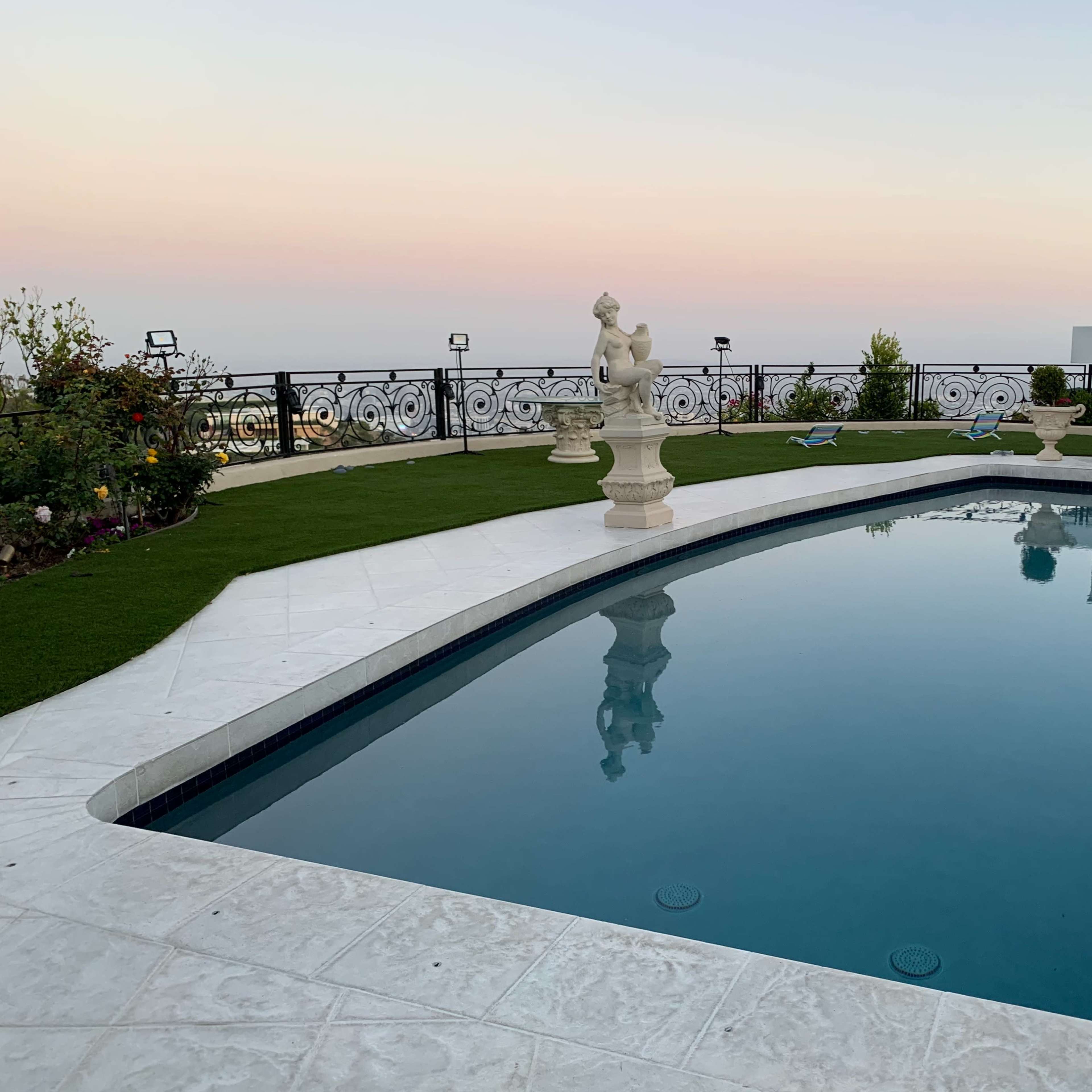 A pool surrounded by a tiled patio, featuring a sculpture and landscaped gardens against a pastel sky.