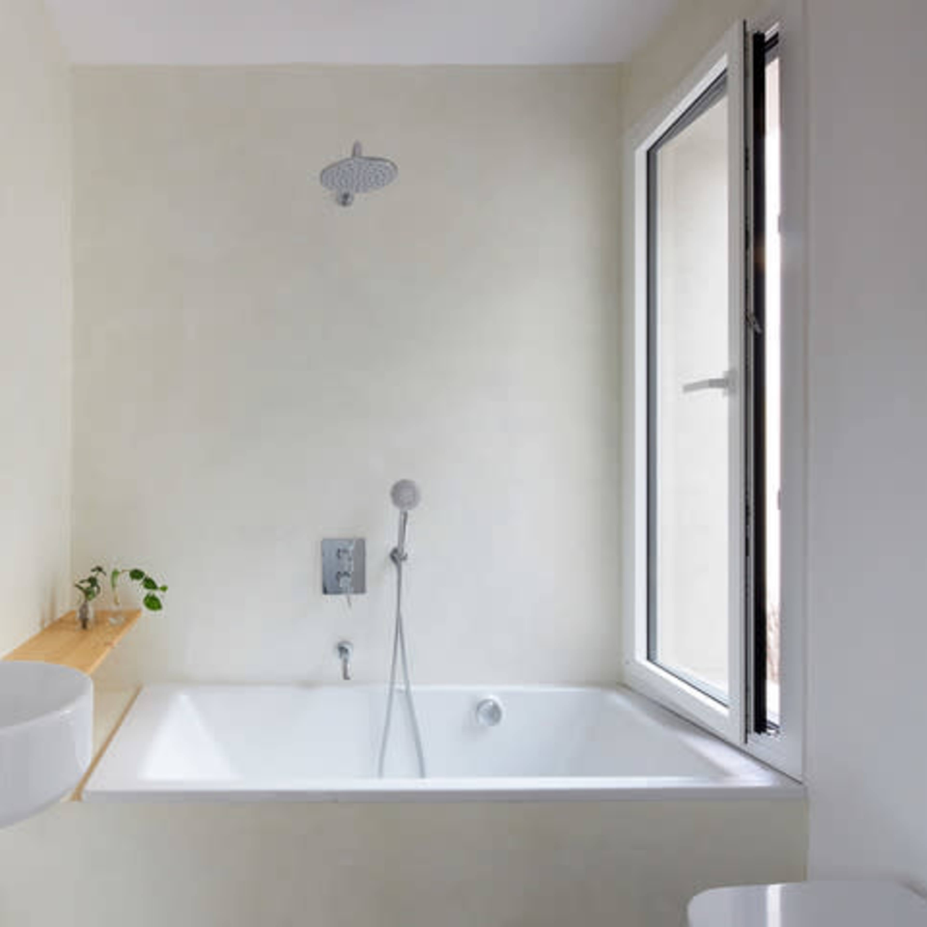 A minimalist bathroom features a bathtub, a wall-mounted sink, and a showerhead, with a small plant on a wooden shelf near a window.