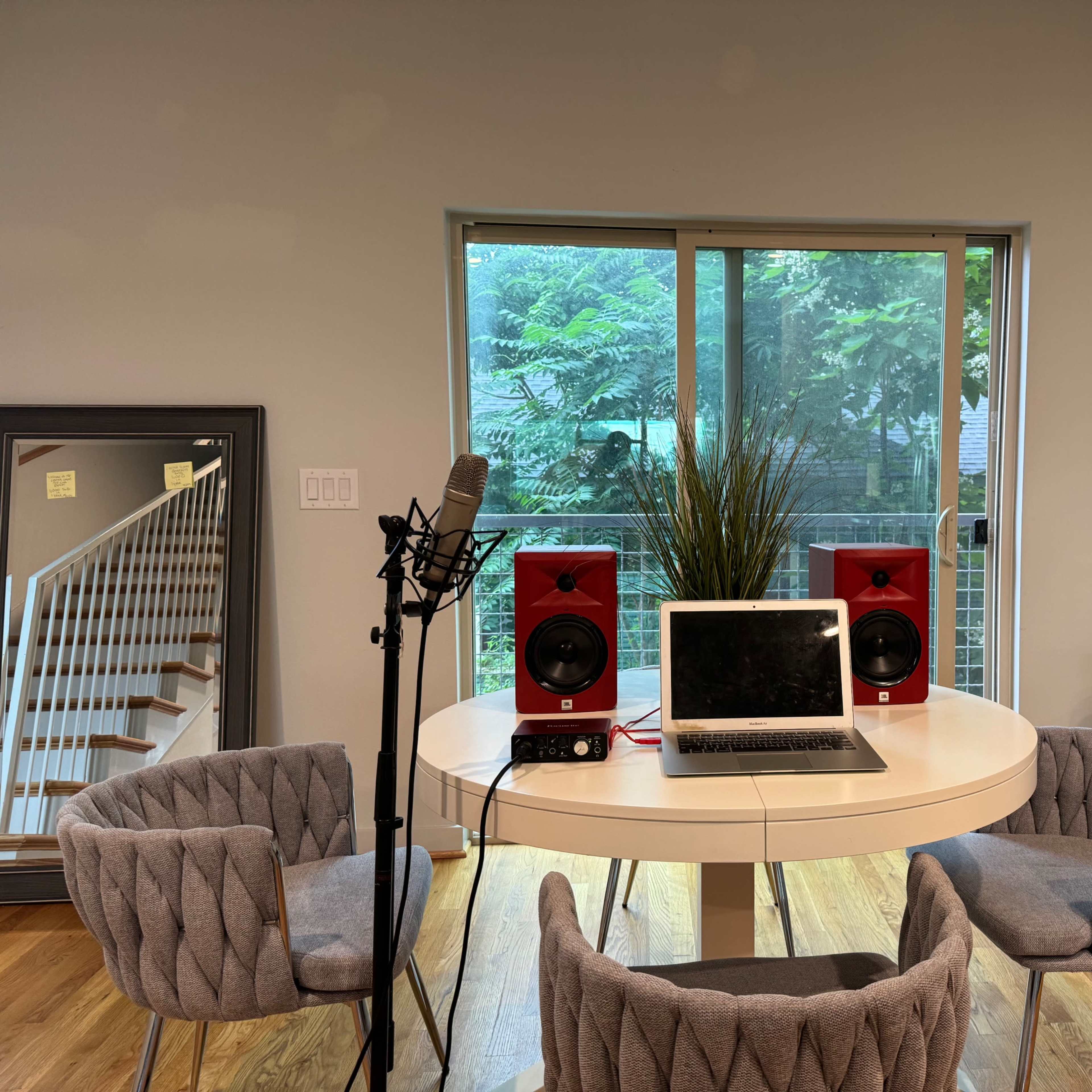 A modern workspace features a round table with a laptop and studio monitors, surrounded by four chairs, next to a large window with greenery outside.