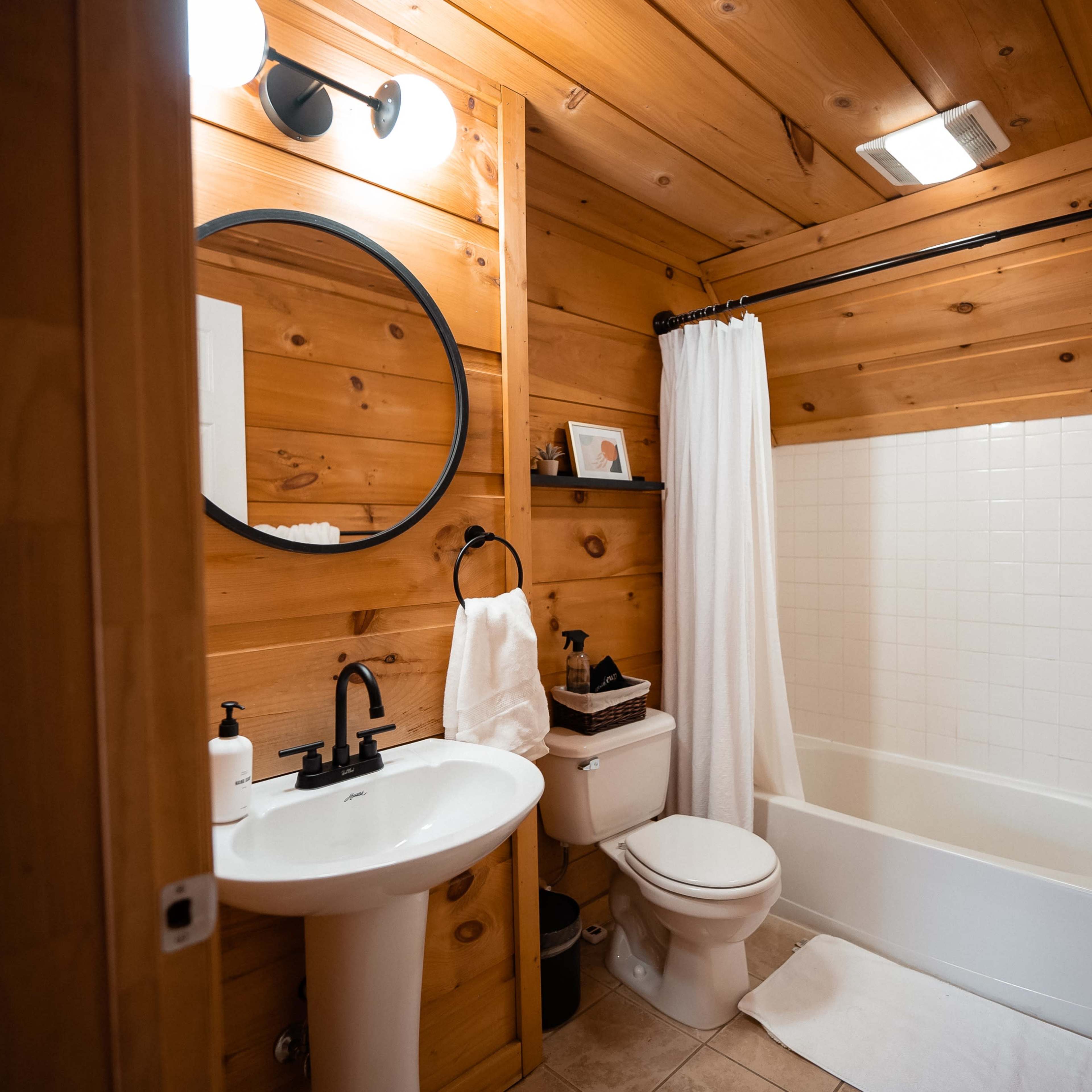 The bathroom features wooden walls, a round mirror above a sink, a white bathtub, and a toilet, with a shower curtain and toiletries on display.