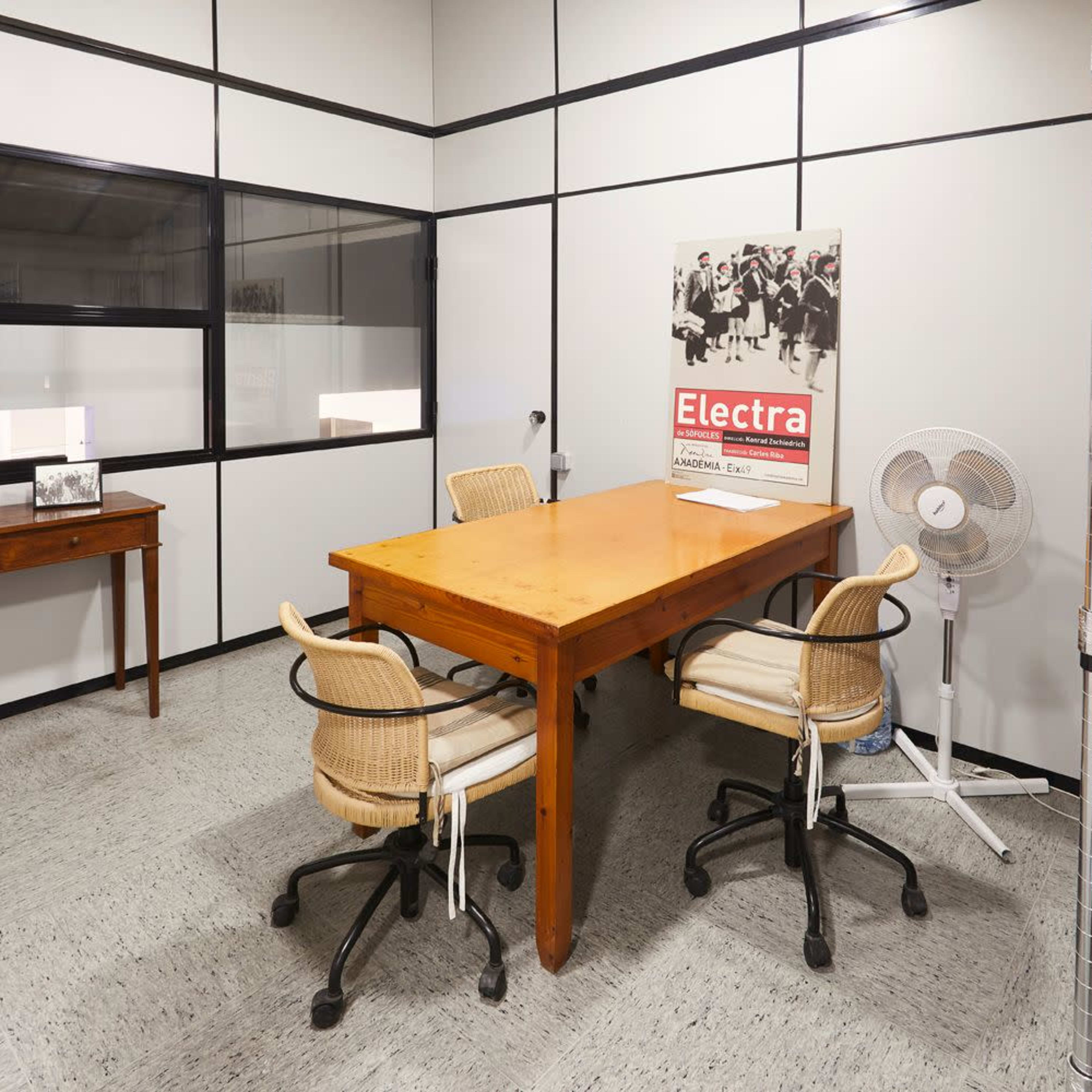 A small office with a wooden table surrounded by four chairs, a wall fan, and a framed photo on a side table.
