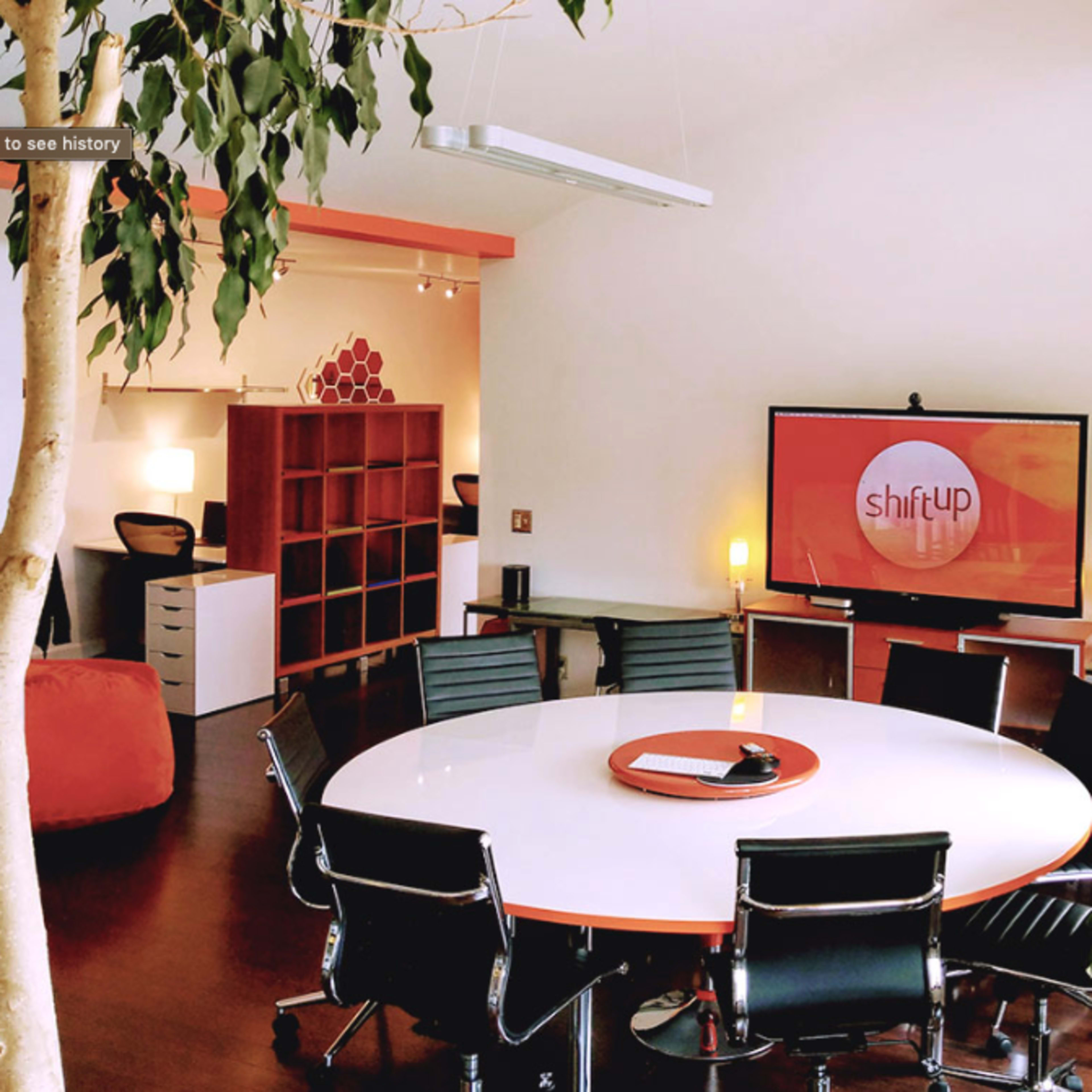 A modern office space features a large round table surrounded by black chairs, with a television displaying the logo "shiftUP" and a tree in the corner.