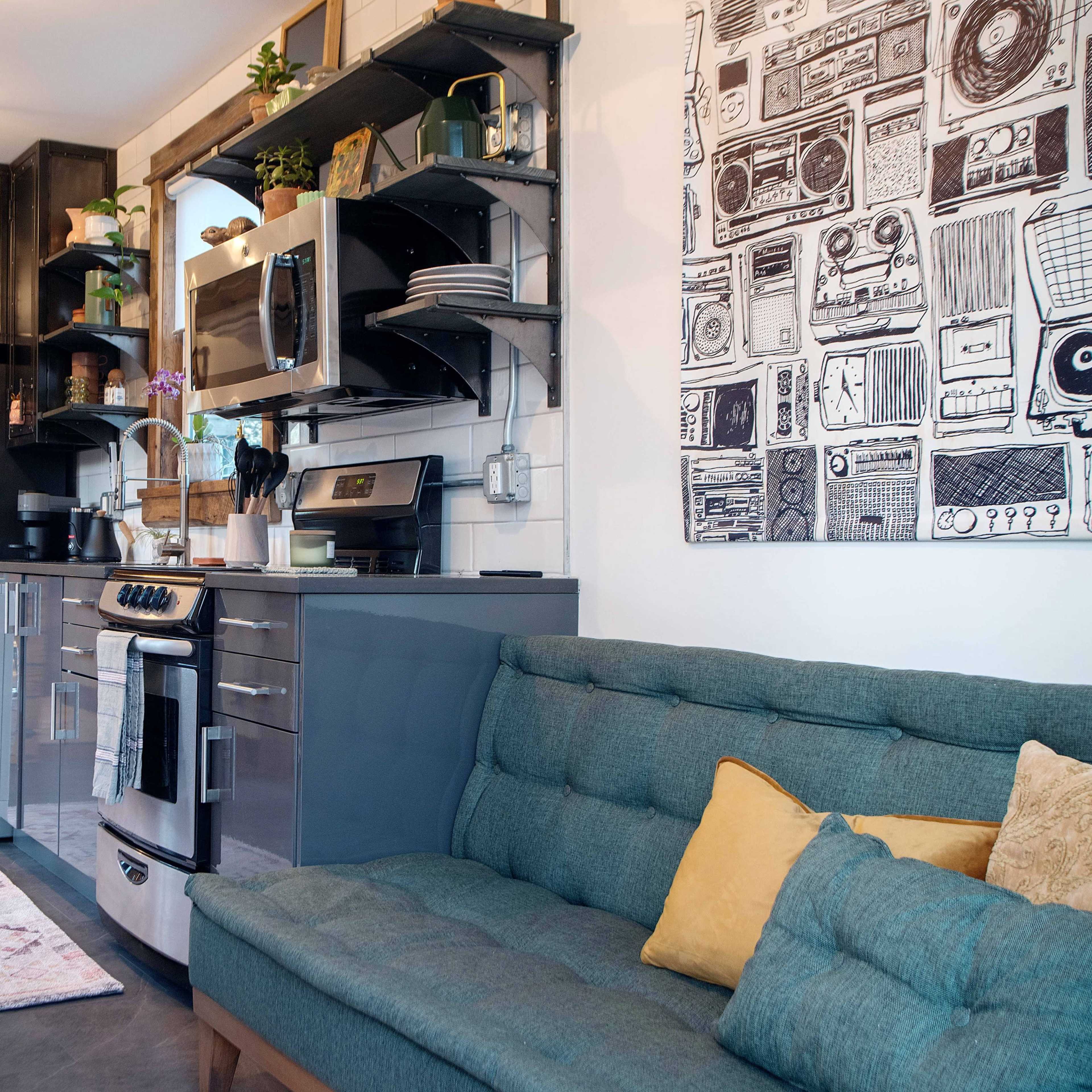 A compact kitchen area with modern appliances, wooden shelves, a sofa, and a decorative wall hanging.