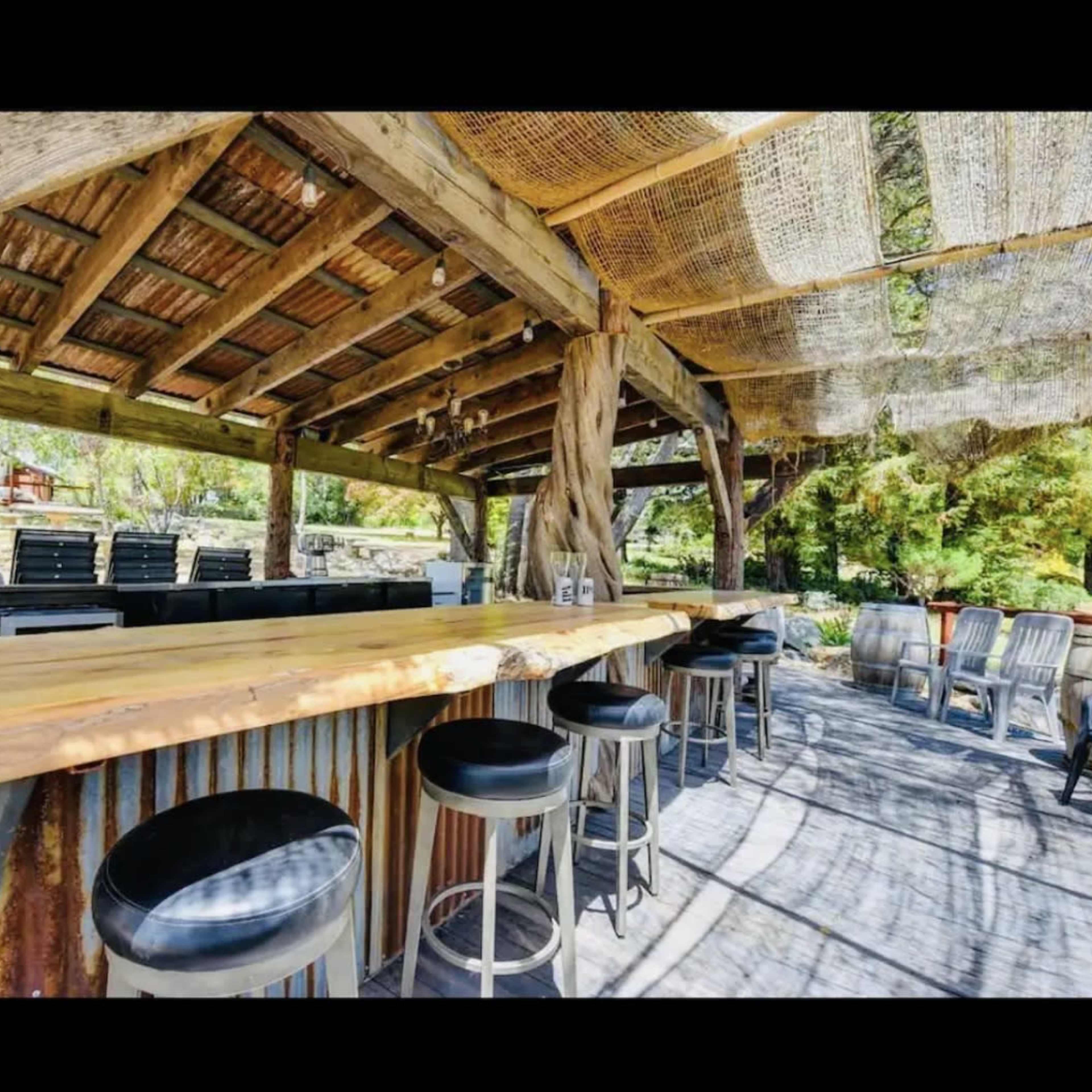 A rustic outdoor bar with a wooden countertop and metal stools is surrounded by a shaded structure made of wood and thatch.