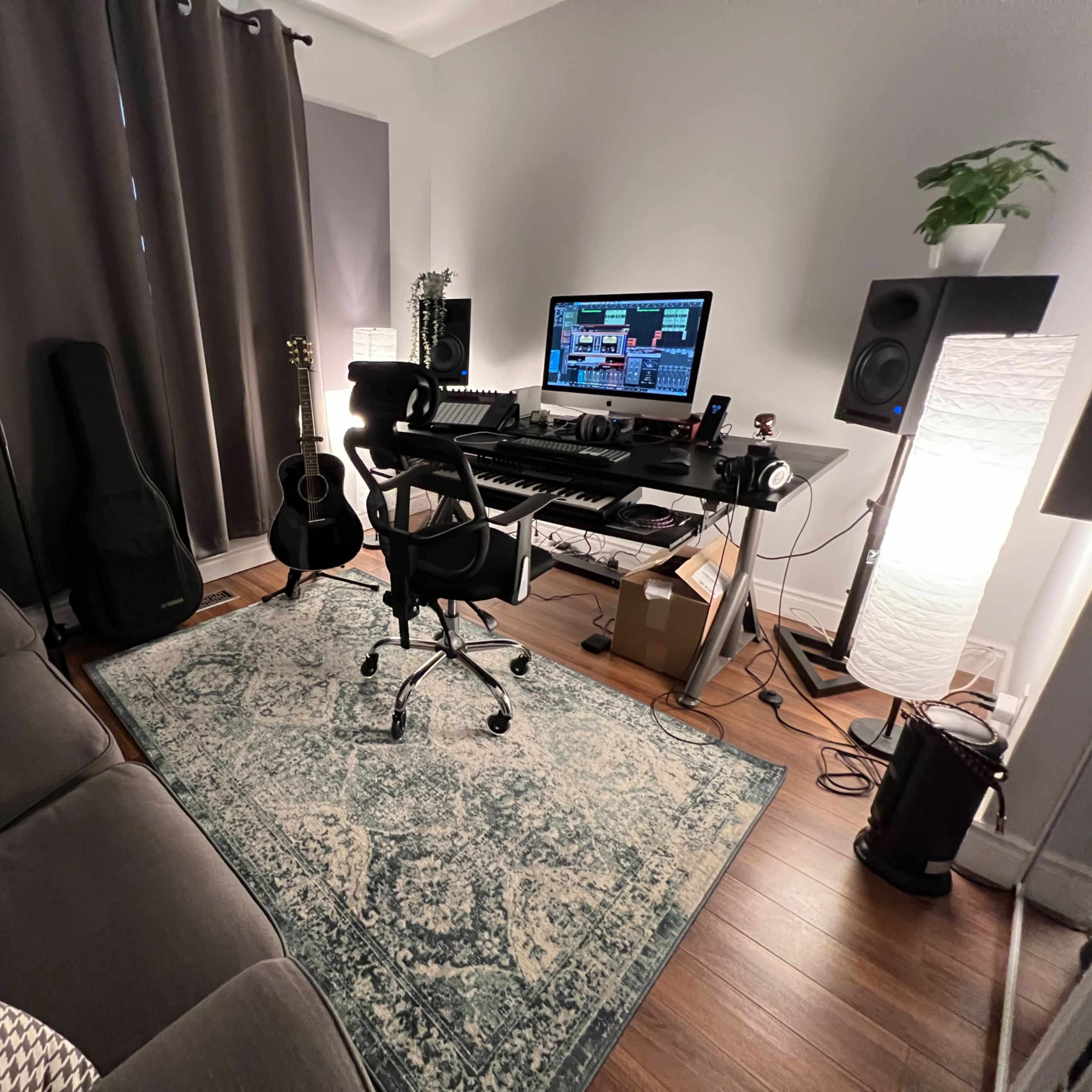 A home music studio featuring a desk with a computer and keyboard, a guitar leaning against a chair, and a cozy sofa with patterned cushions.