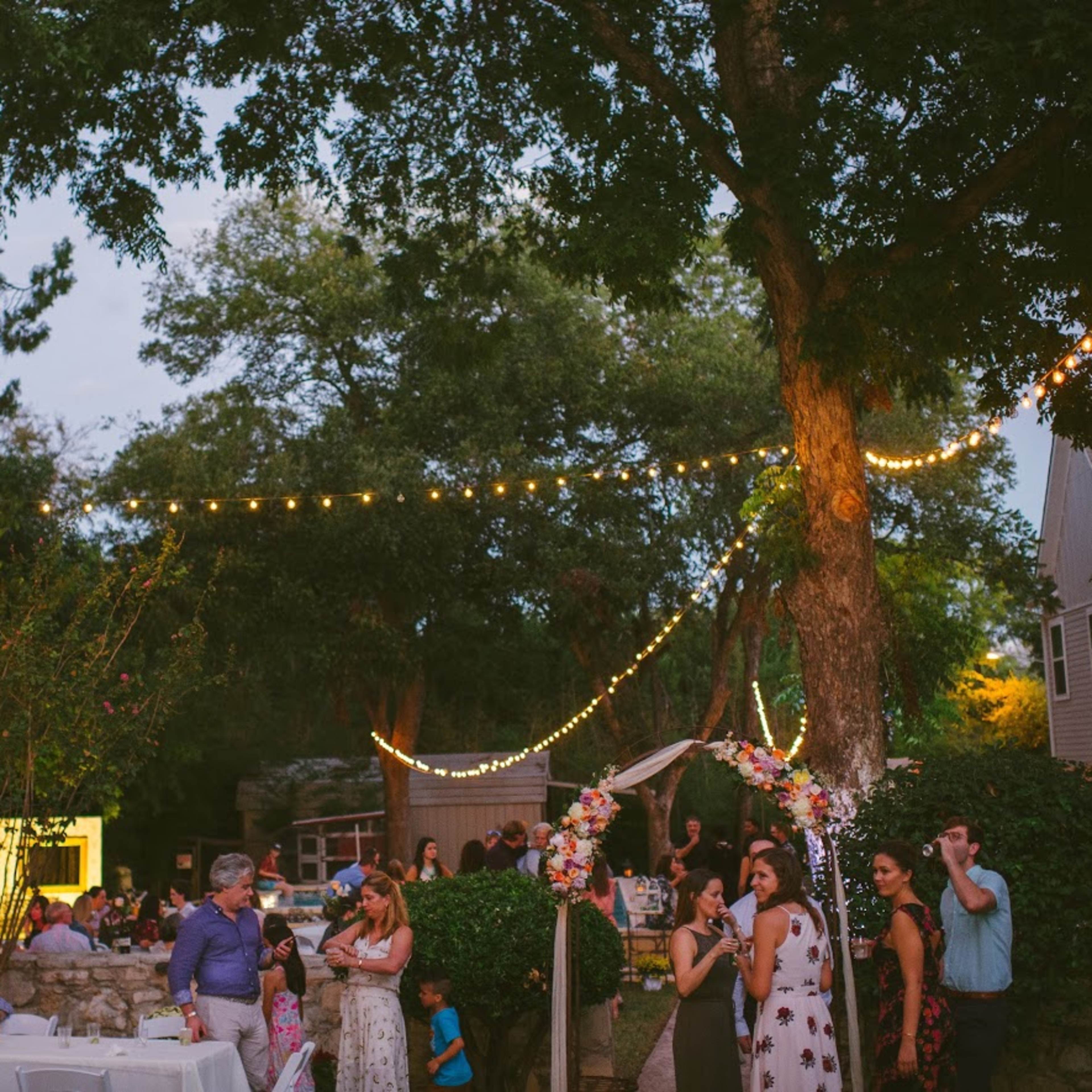 A gathering takes place outdoors in a backyard setting, with string lights overhead and guests mingling among tables and decorations.