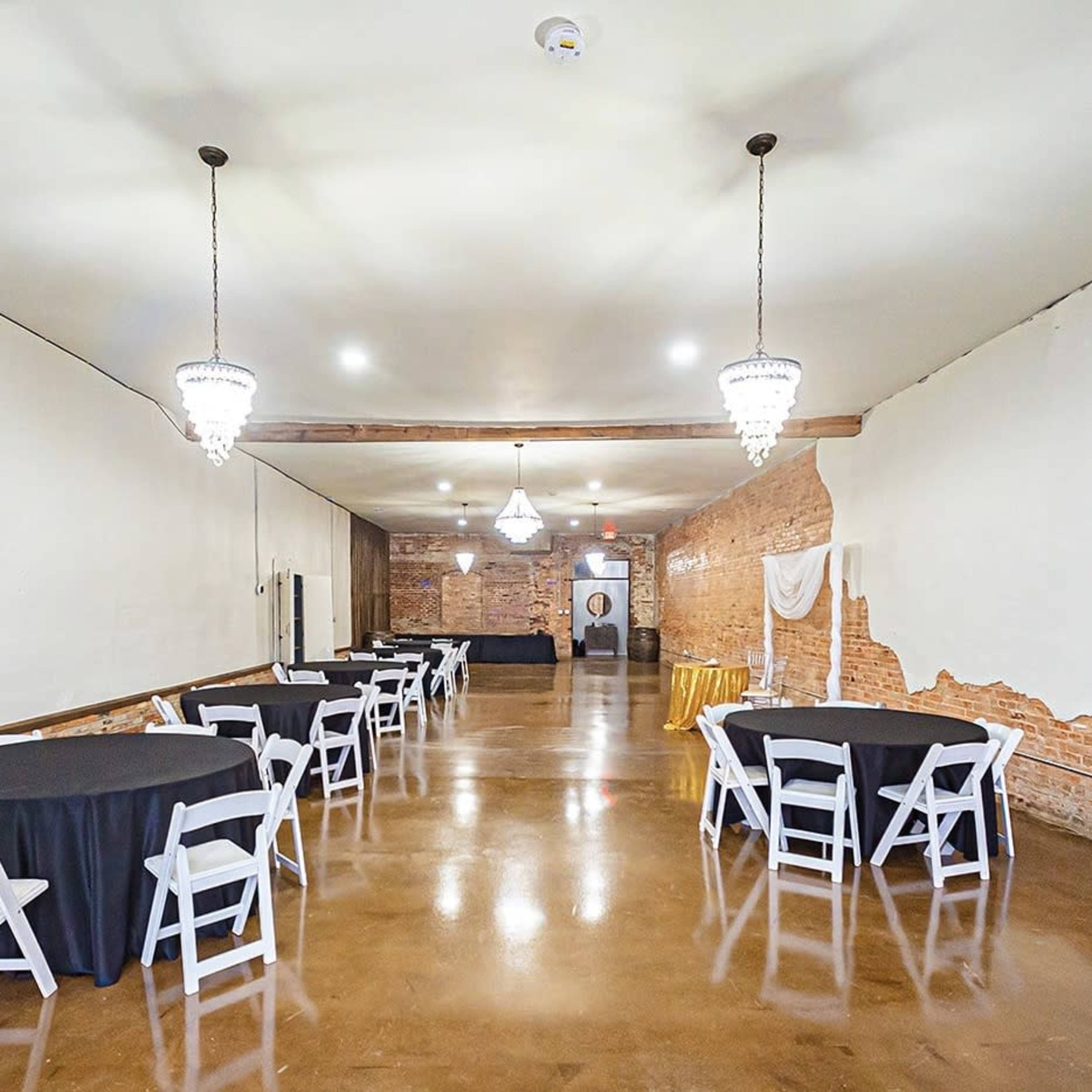 A spacious event venue features round tables with black tablecloths and pendant lights hanging from the ceiling, set against a backdrop of exposed brick walls.