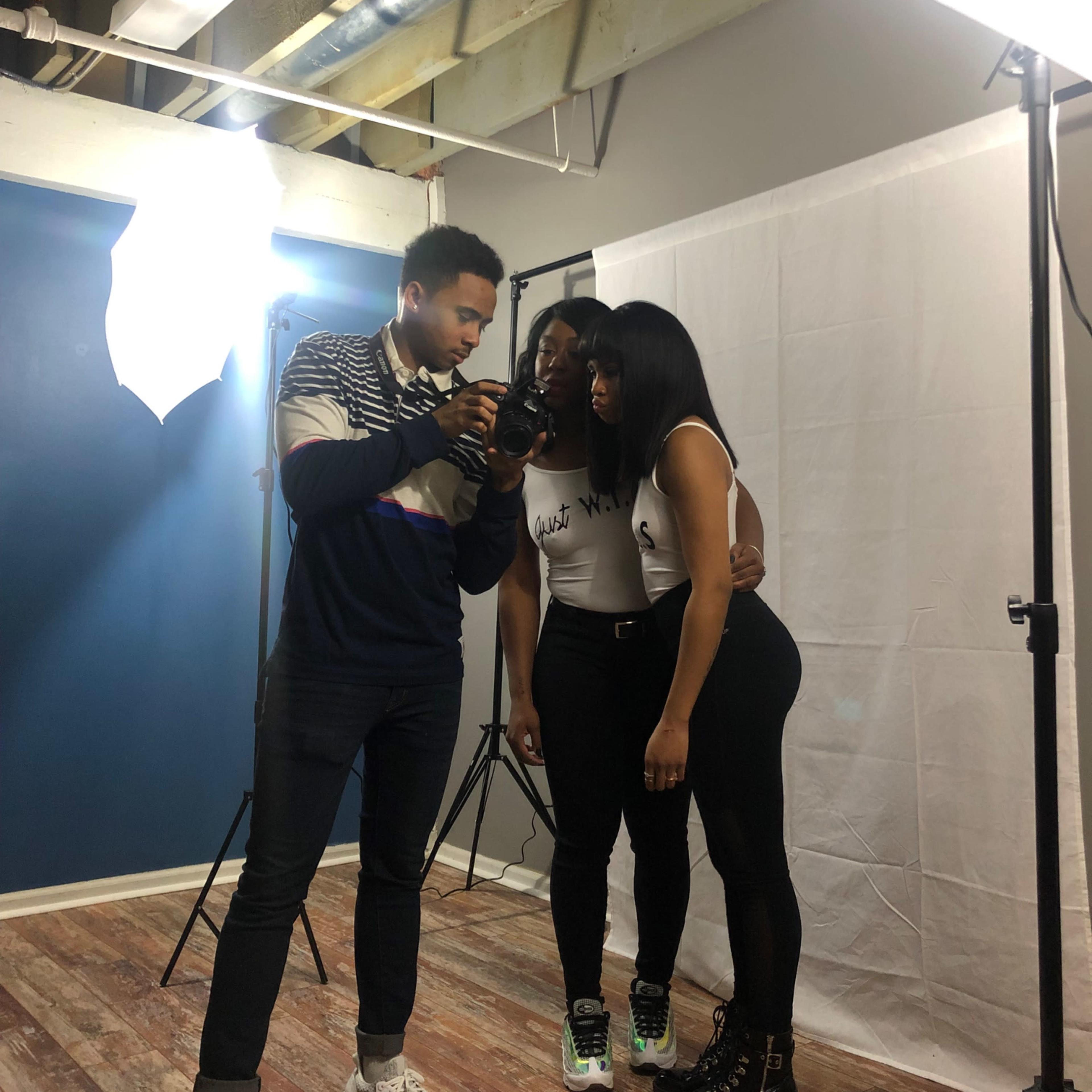 A photographer adjusts his camera while two models pose for a photoshoot in a studio setting.