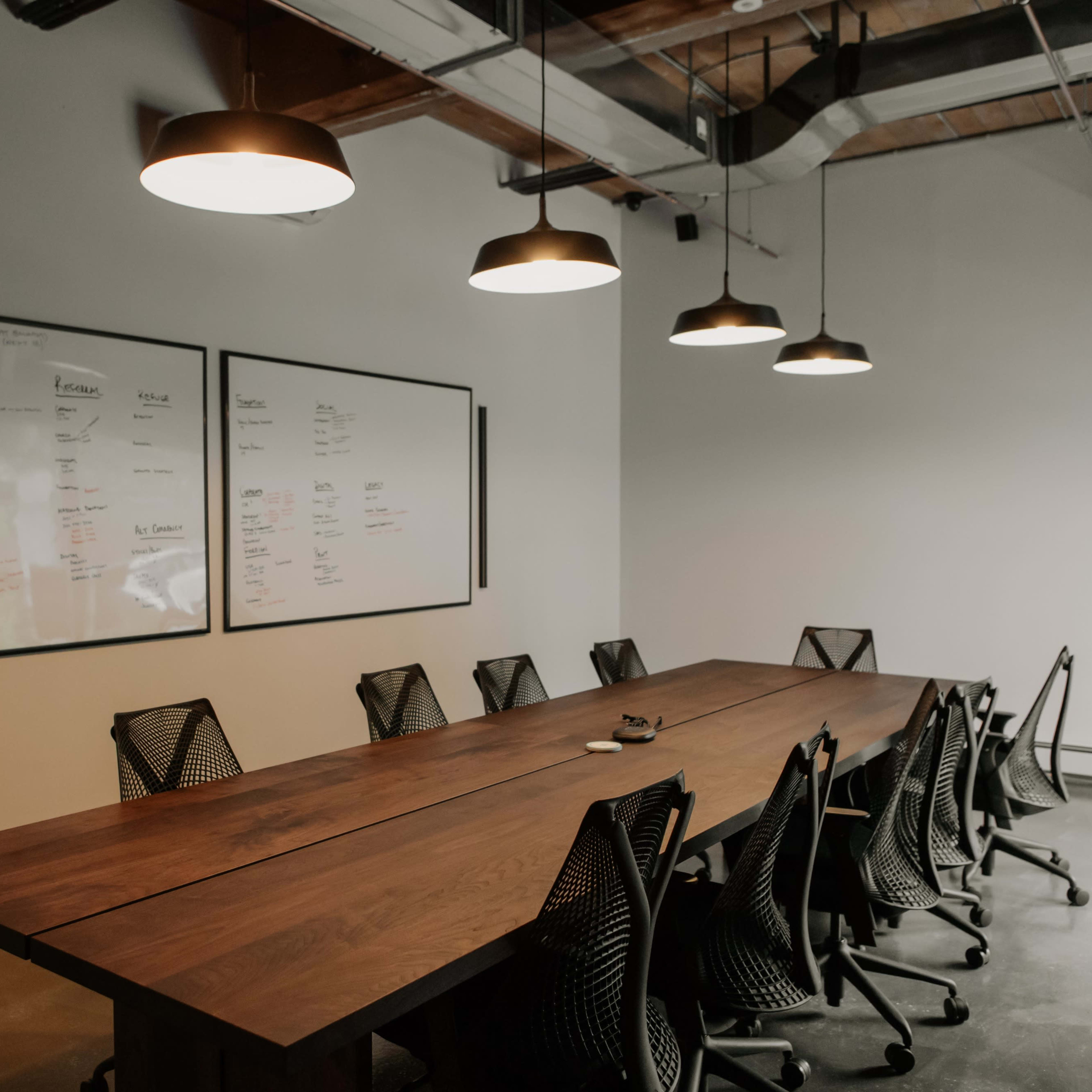 A conference room features a long wooden table surrounded by black mesh chairs and two large whiteboards mounted on the wall.