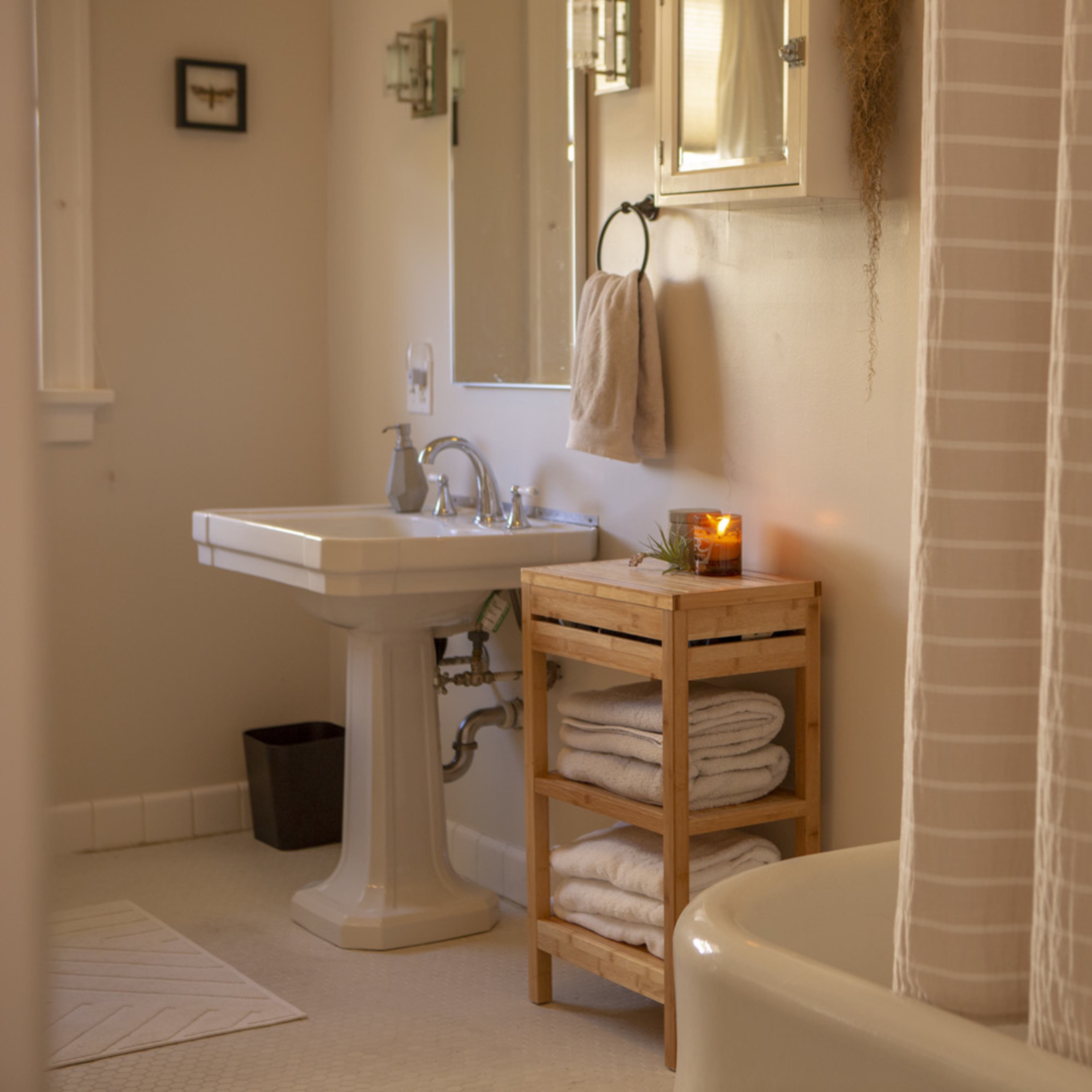 A clean bathroom features a sink, a wooden shelf stacked with neatly rolled towels, and a bathtub partially obscured by a shower curtain.
