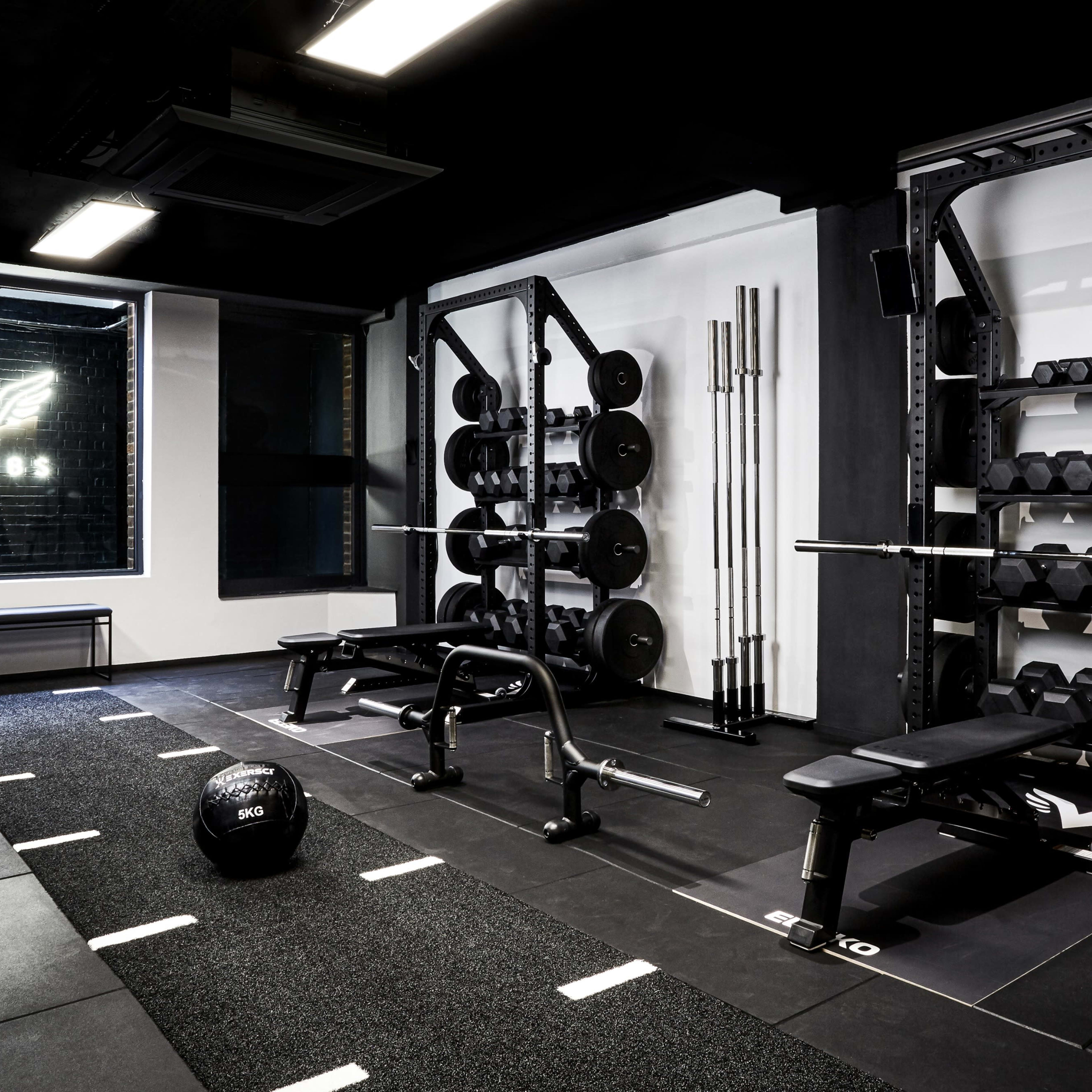A modern gym interior featuring weightlifting equipment, including benches, dumbbells, and a barbell rack, set against a backdrop with a logo.