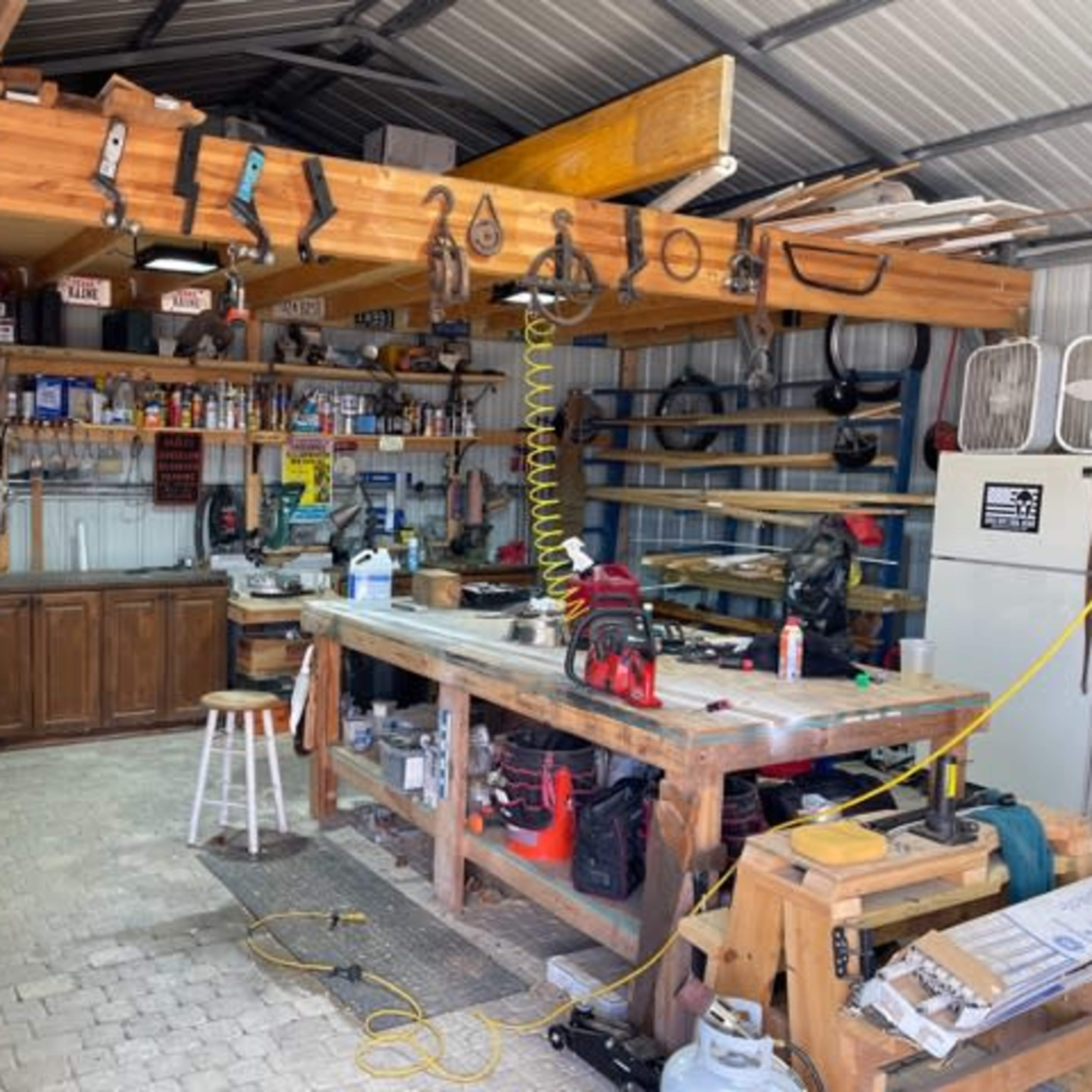 A cluttered workshop with various tools and equipment arranged on shelves and counters beneath a metal roof.