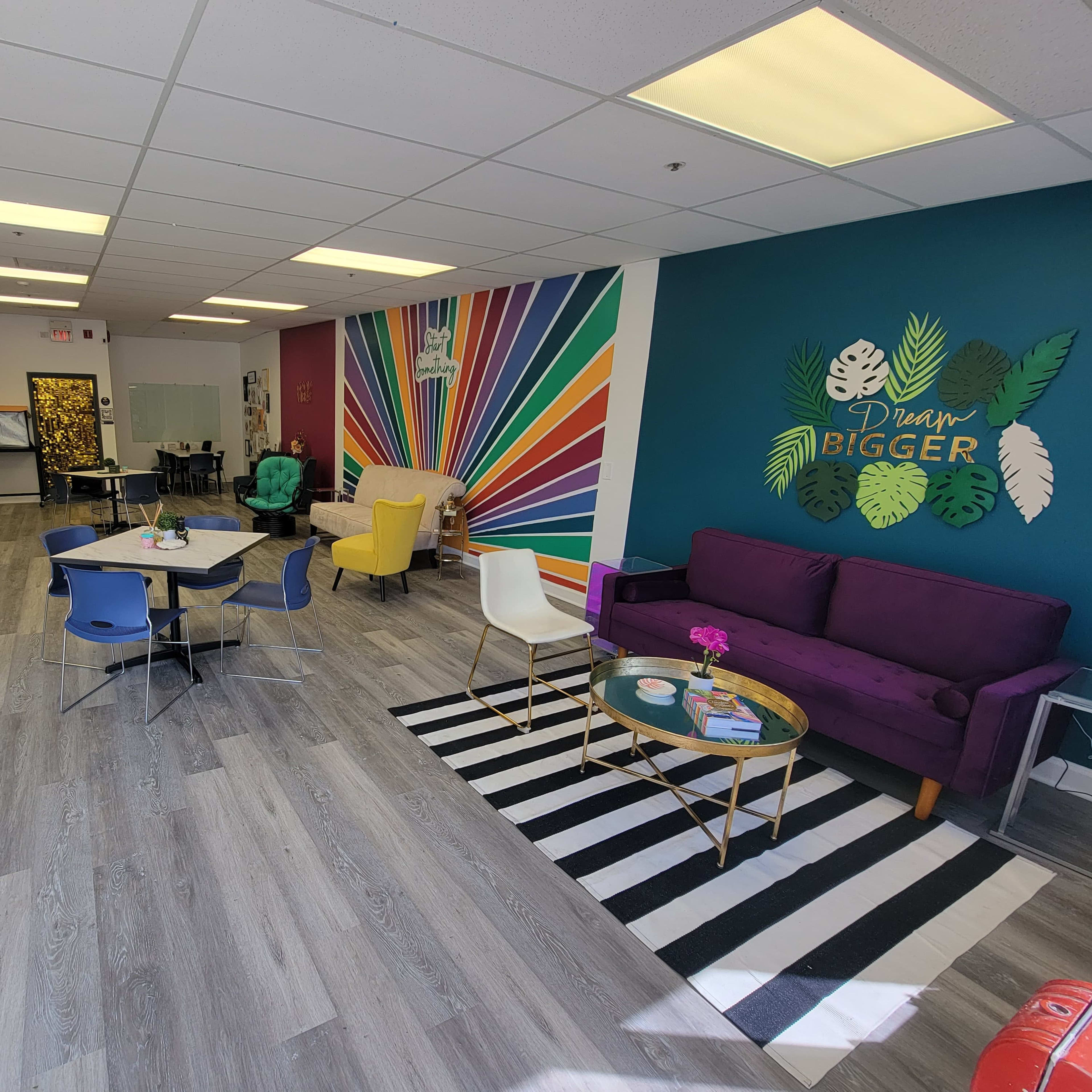 A brightly colored, modern open space featuring a seating area with a purple sofa, tables, and vibrant wall art.