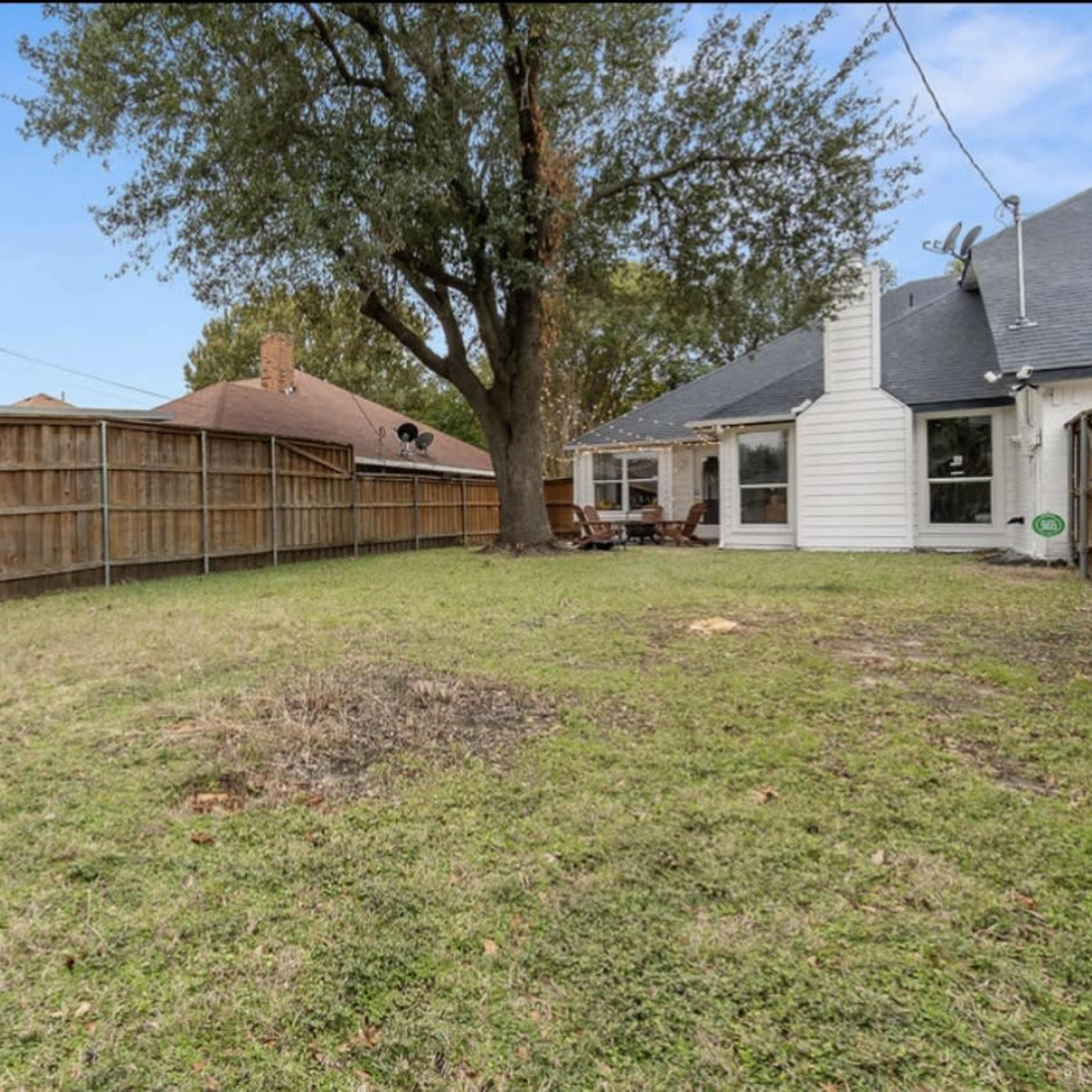 A fenced backyard with grass, a large tree, and a house with a patio area on one side.