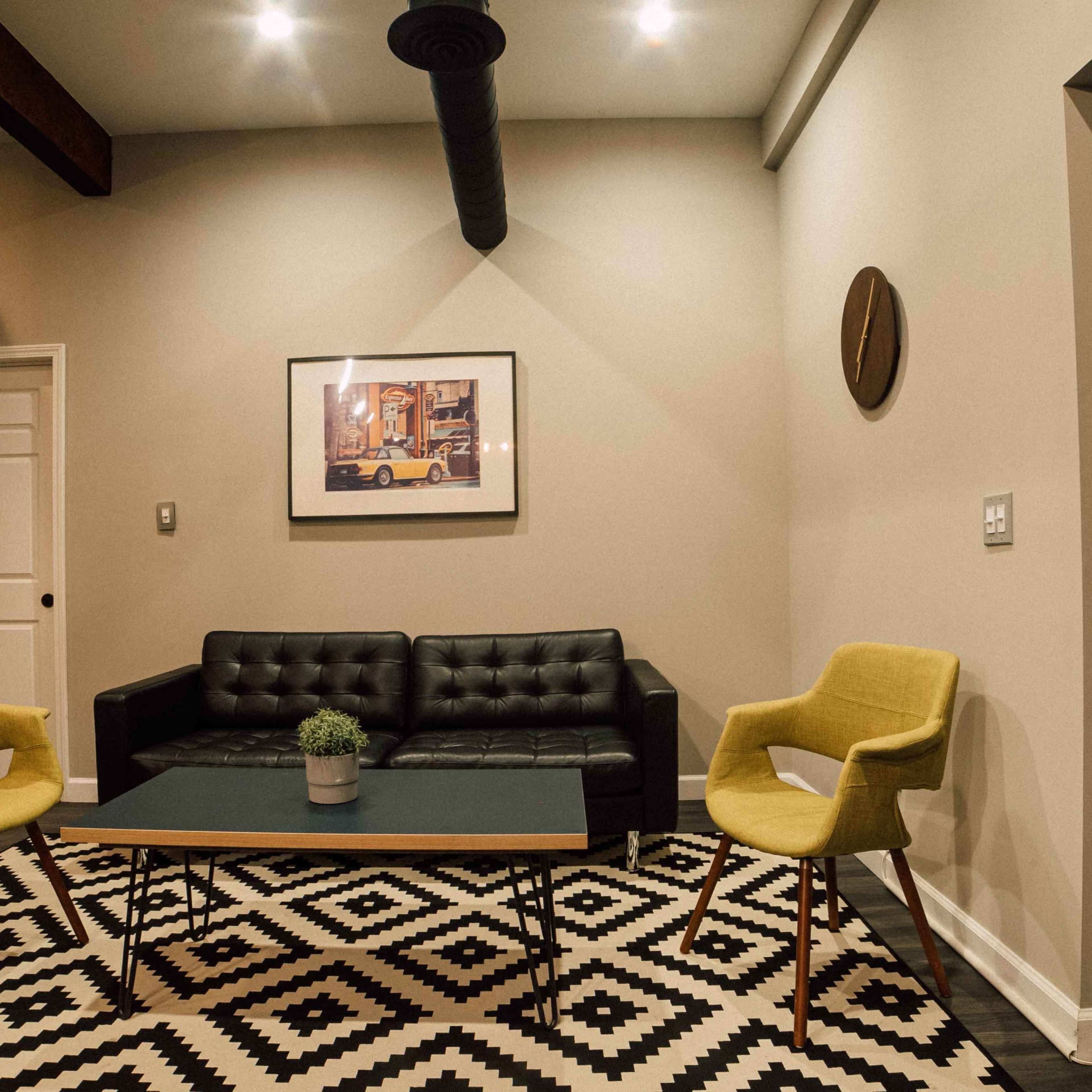 A modern sitting area with a black couch, two yellow chairs, a coffee table with a potted plant, and a geometric patterned rug.