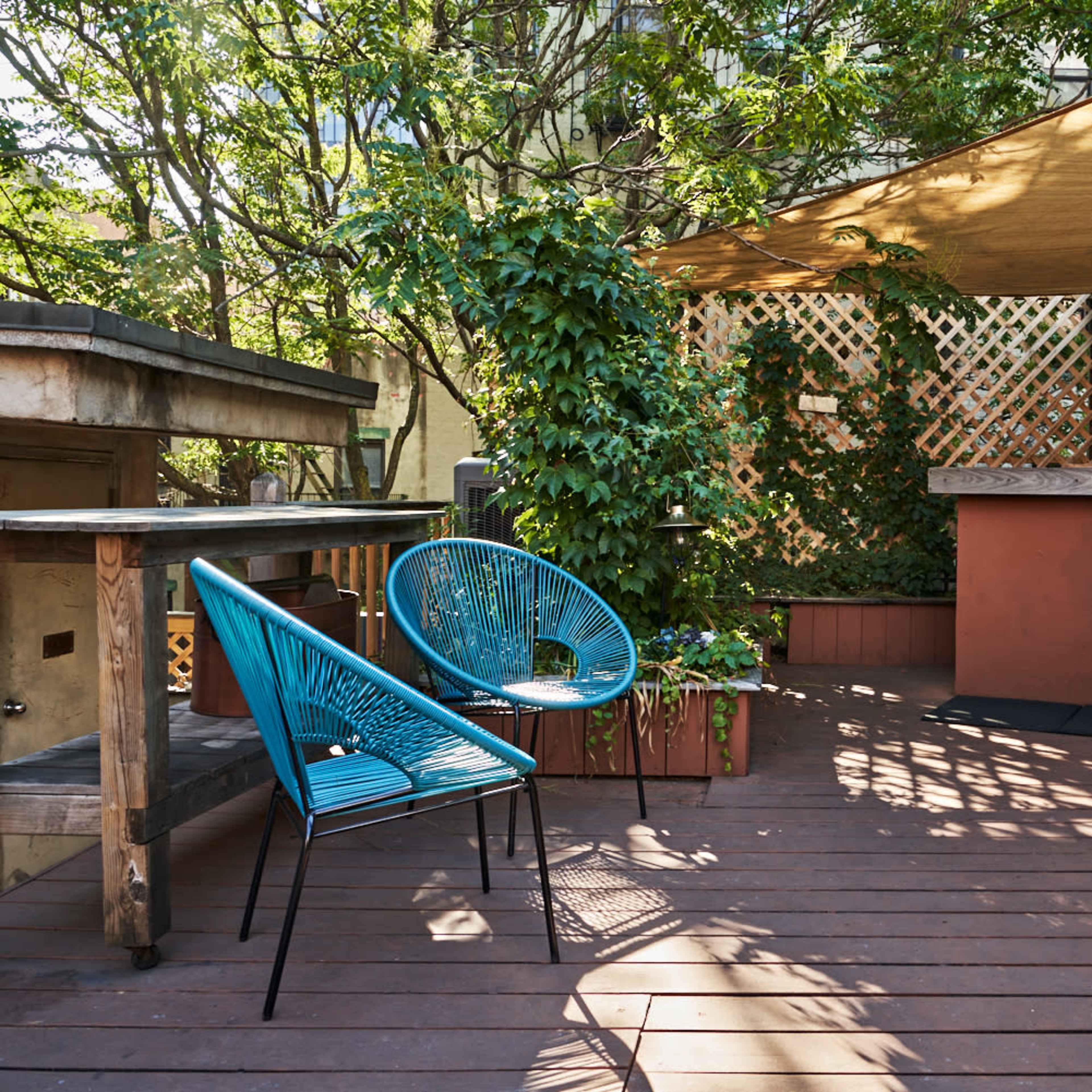 A patio with wooden flooring, a bar area, an outdoor cooking station, and blue chairs surrounded by greenery and a canopy.