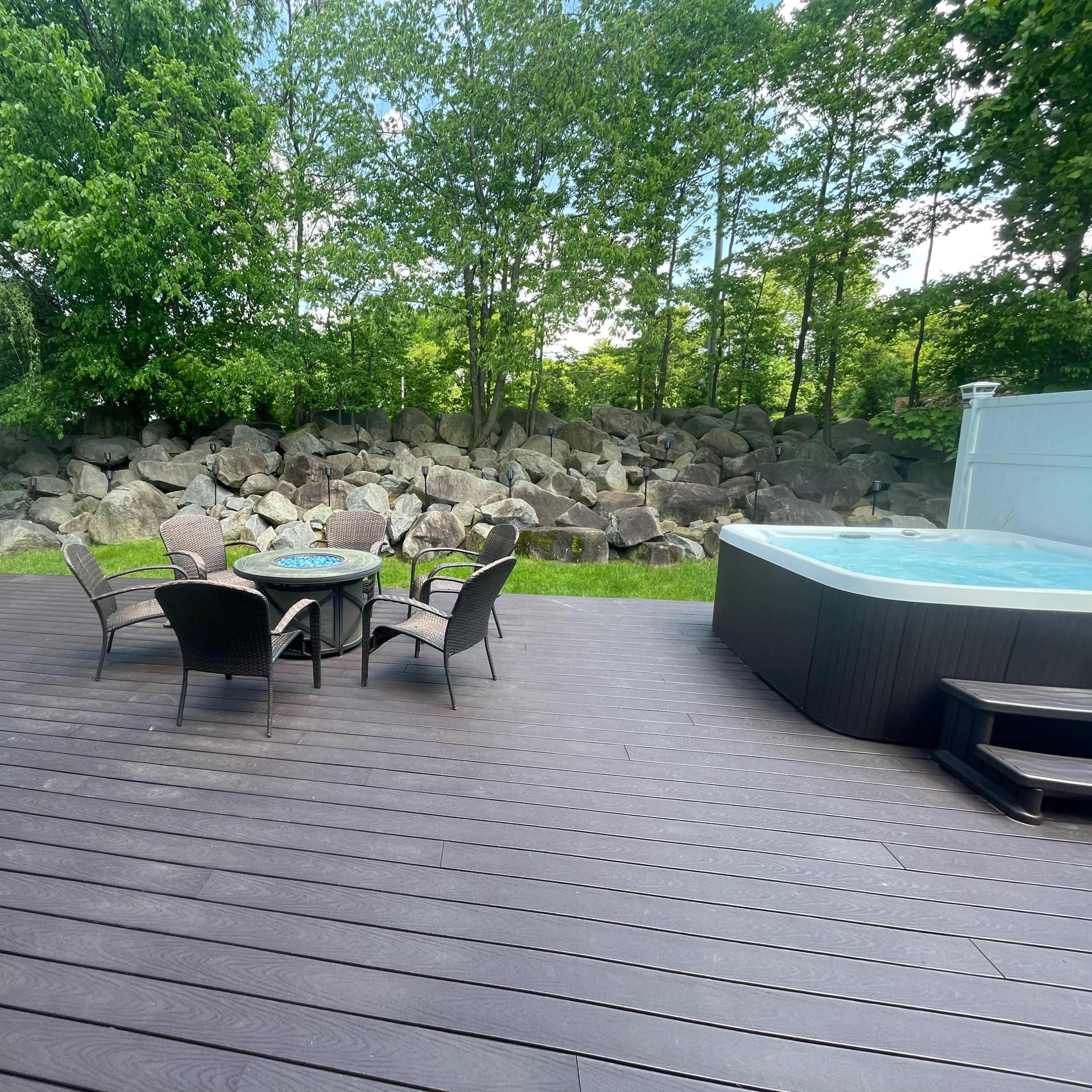 A wooden deck with a seating area featuring a table and chairs, alongside a hot tub and a backdrop of large rocks and greenery.