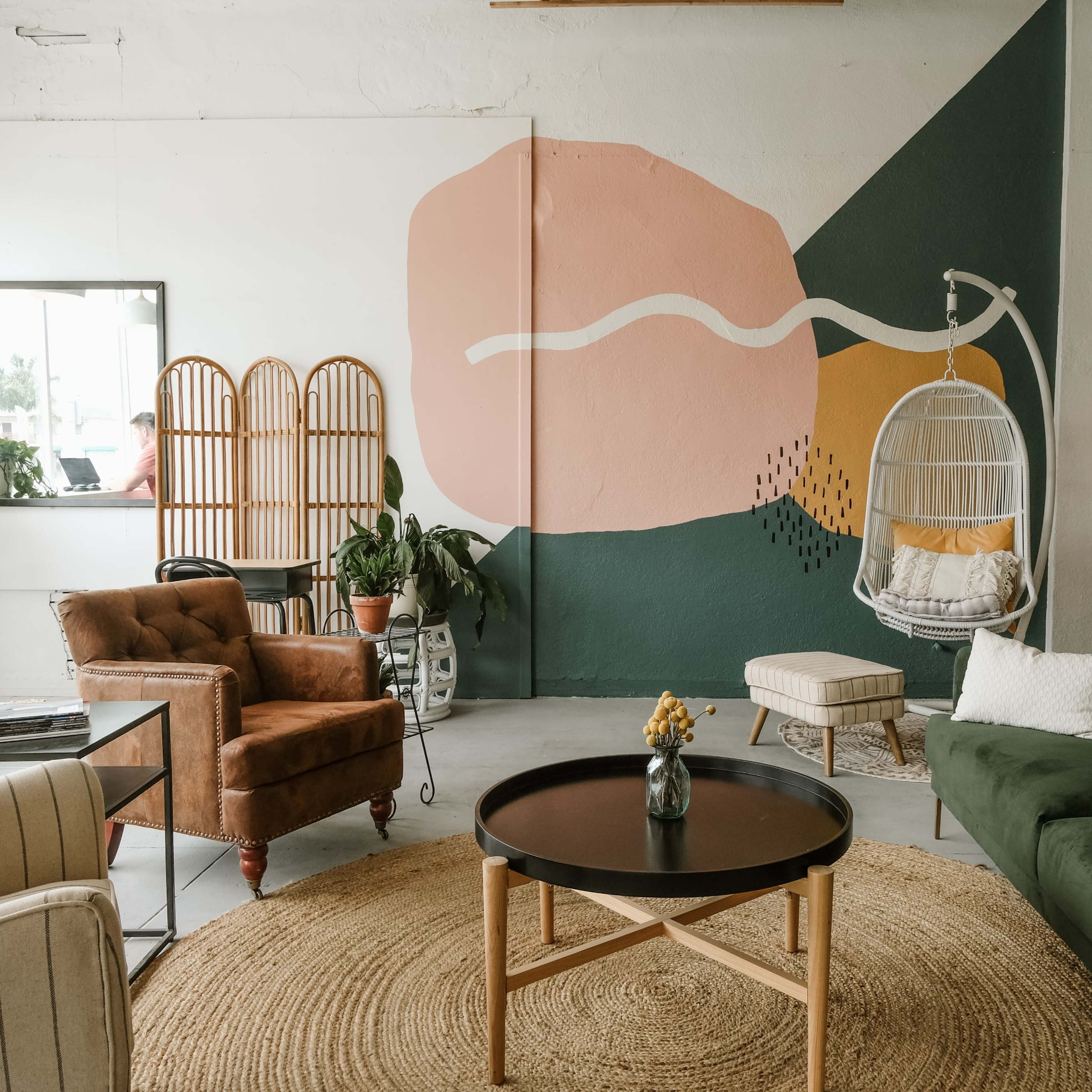 A modern, airy interior space with a green sofa, a hanging chair, a round table, and an artistic wall mural in soft colors.