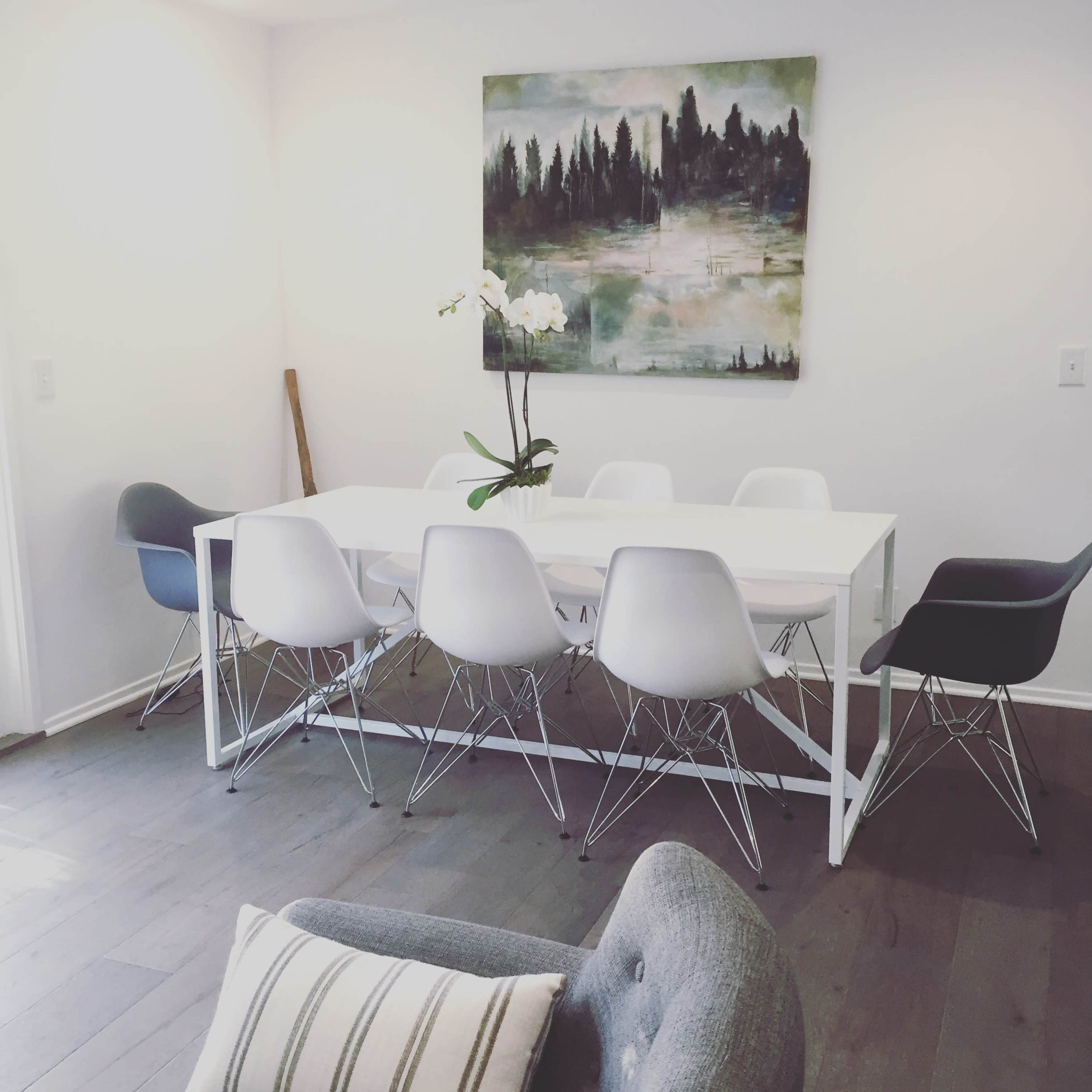 A modern dining area features a white table surrounded by six chairs of varying colors, with a large painting of a forest landscape on the wall.