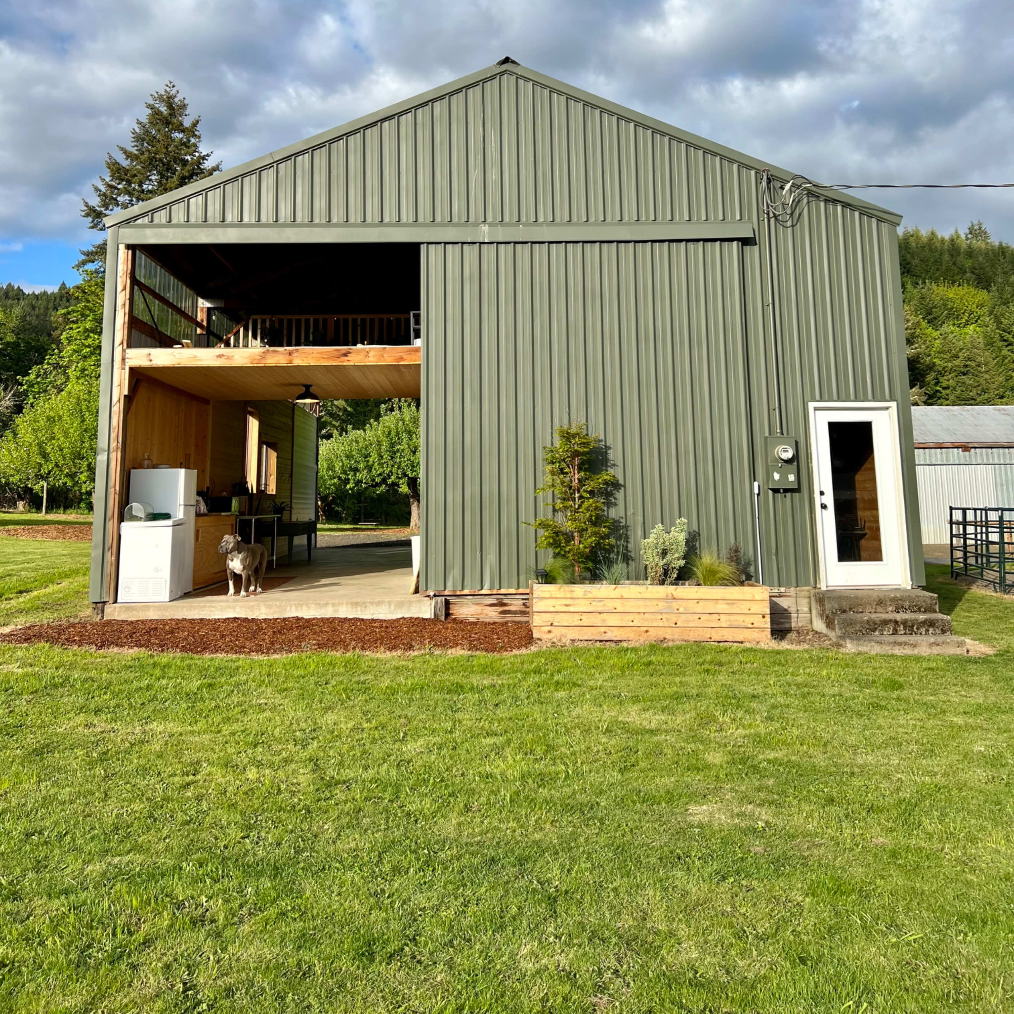 A large green metal barn features an open front with a wooden deck, set on a grassy landscape under a partly cloudy sky.