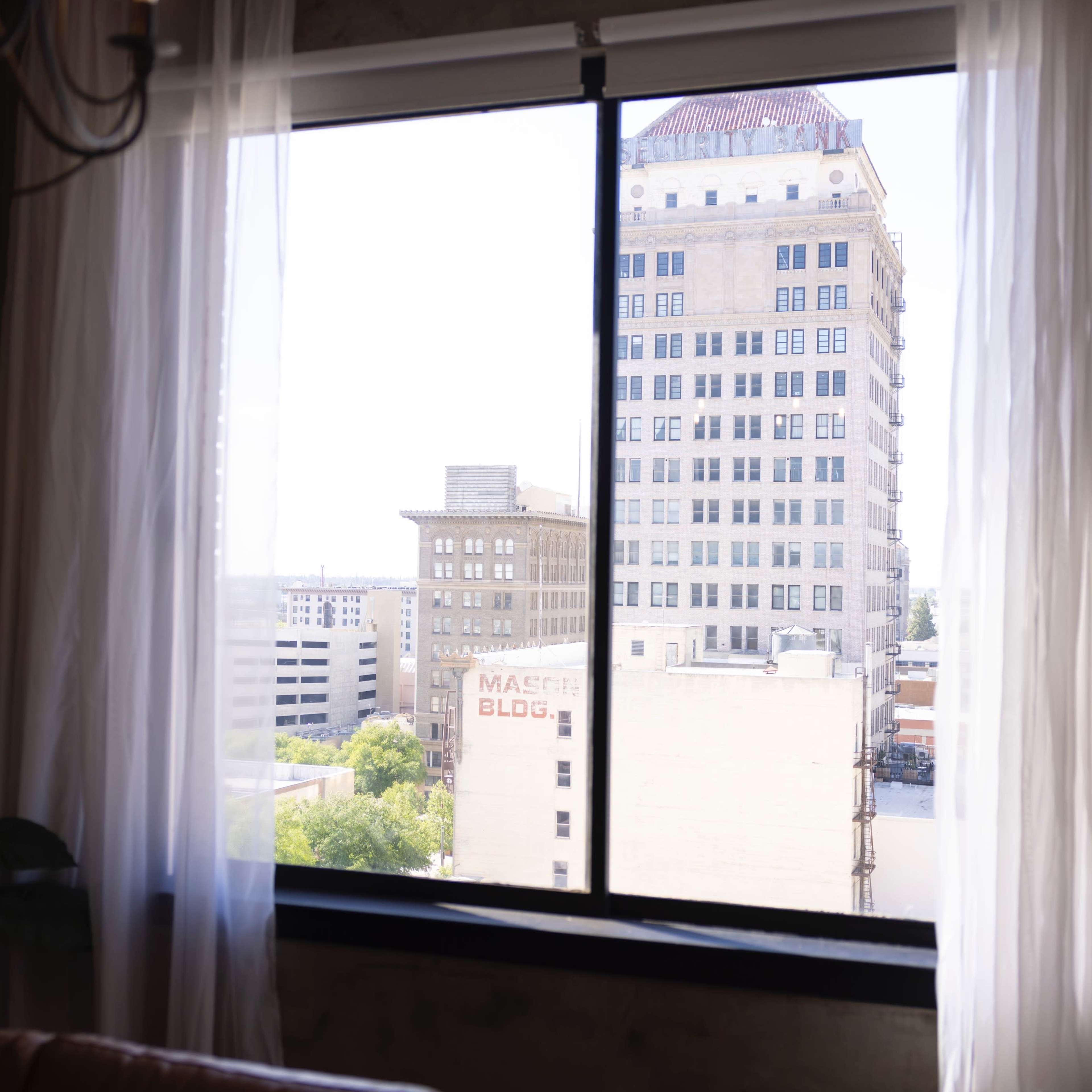 A view from a window with sheer curtains, overlooking a cityscape that includes several buildings.