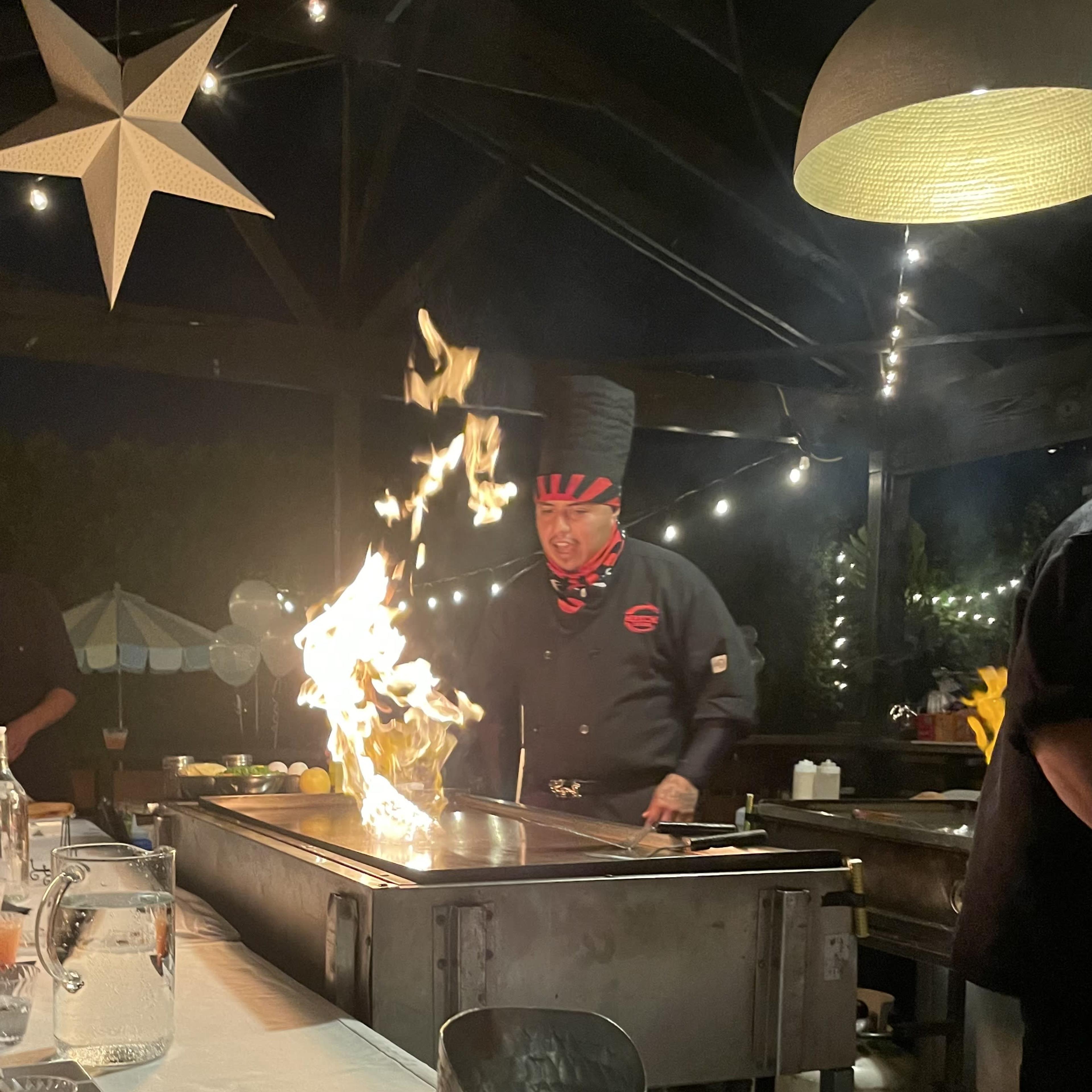 A chef with a tall hat stands at a grill, producing flames while cooking in a dimly-lit outdoor setting adorned with string lights.