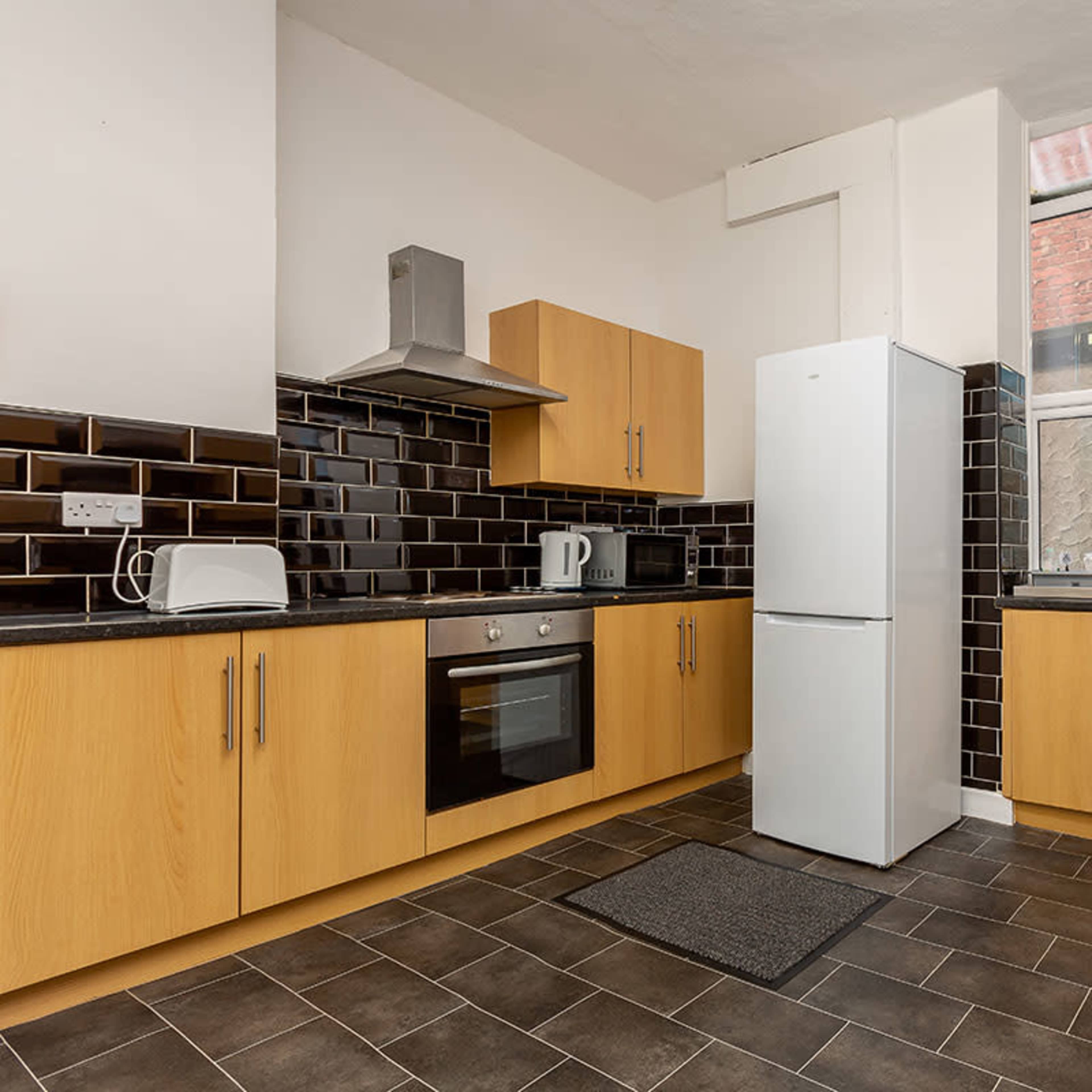 A modern kitchen features wooden cabinetry, a white refrigerator, a stainless steel oven, and dark tile flooring, illuminated by natural light from a large window.