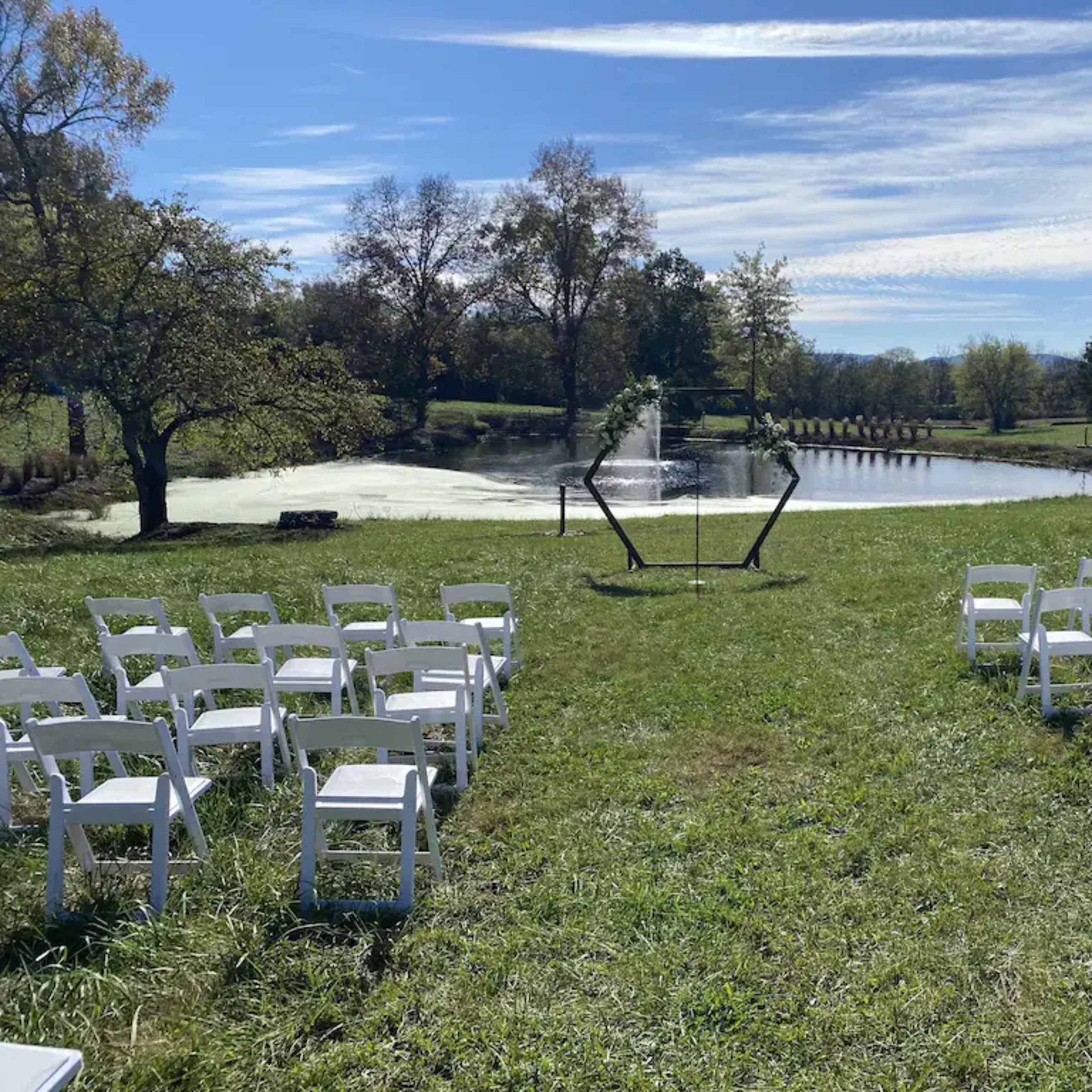 A wedding setup with white chairs arranged in rows facing a small pond surrounded by greenery.