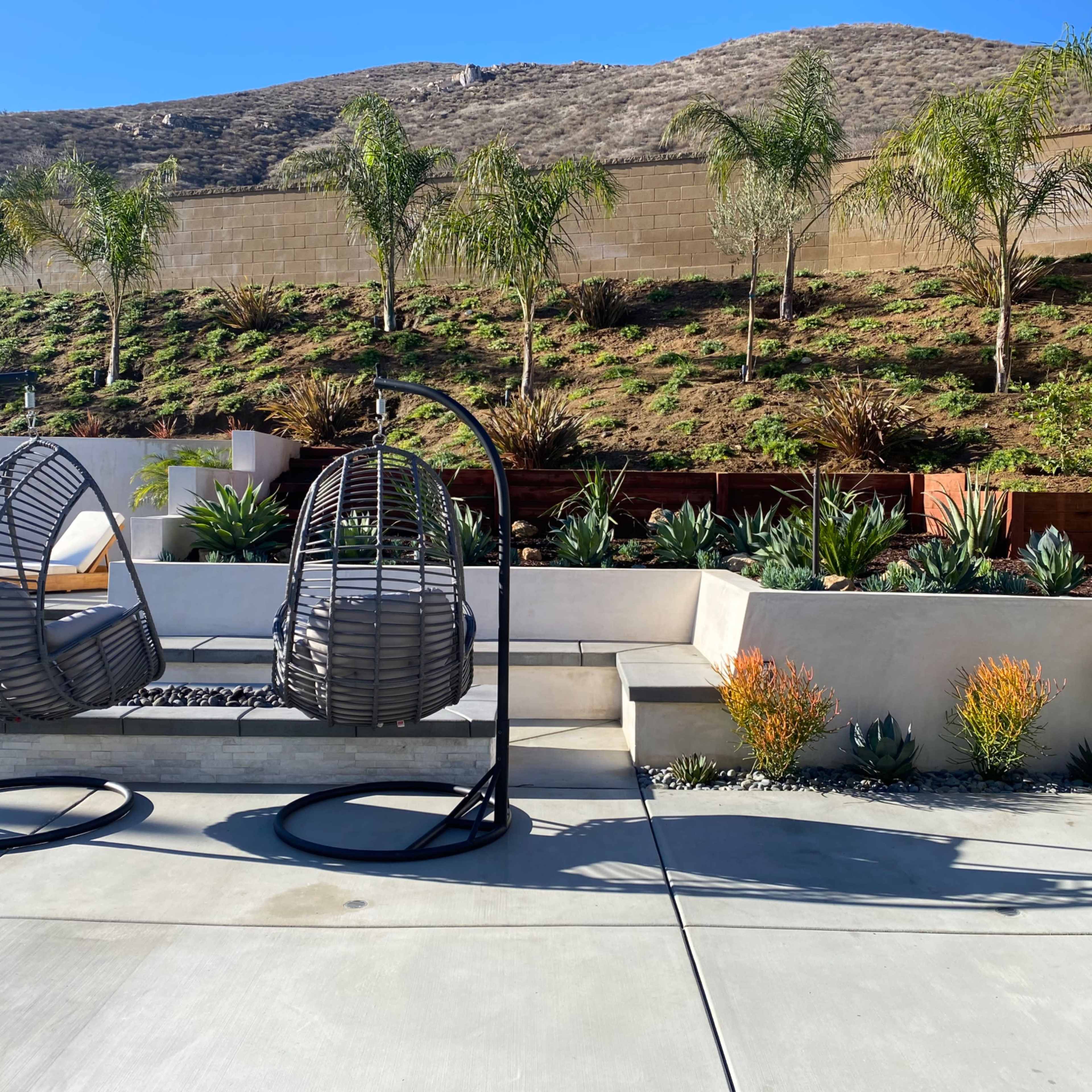 A backyard with two hanging chairs, a tiered landscape featuring plants and shrubs, and a concrete patio.