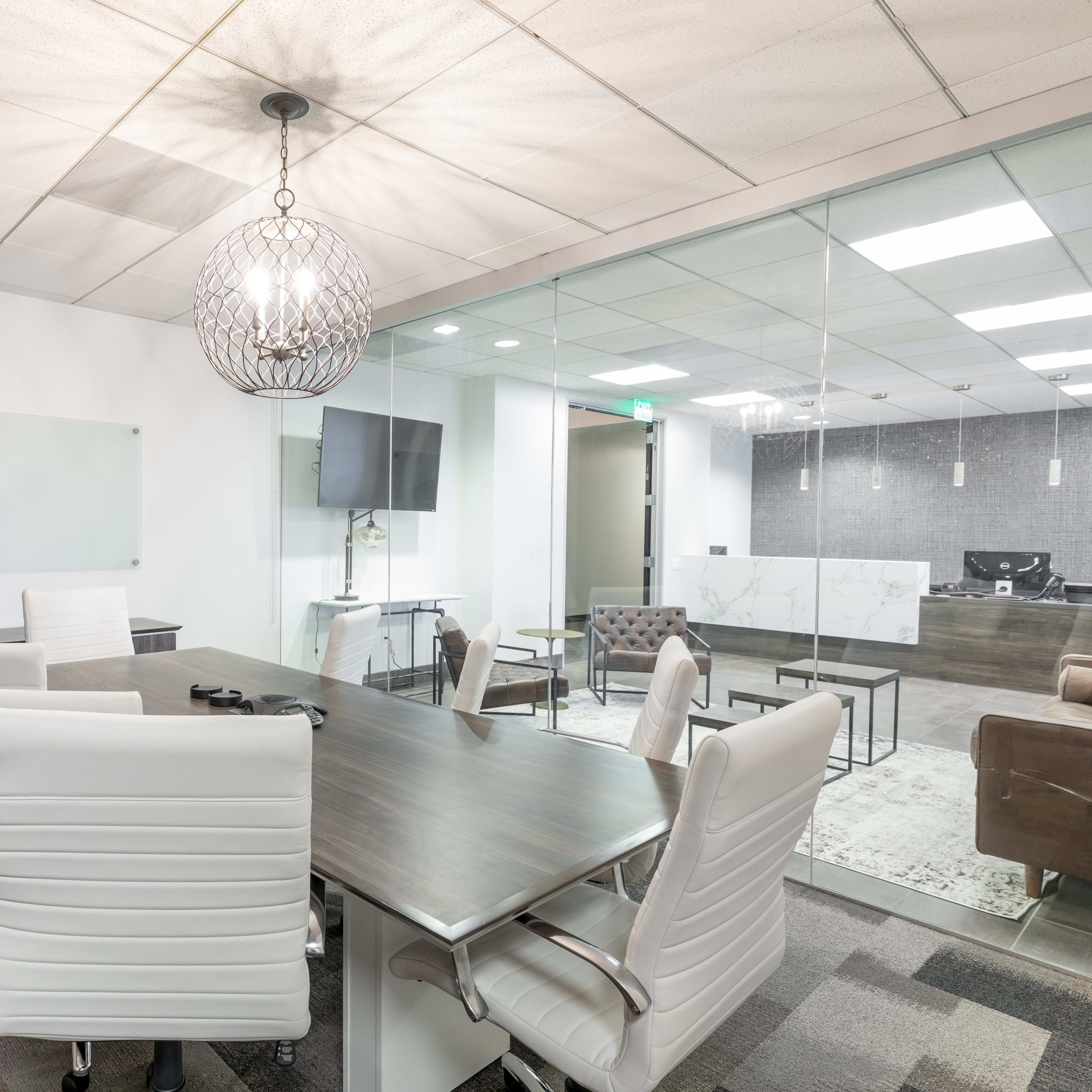 A modern conference room features a large table surrounded by white chairs, with a glass wall leading to an adjoining office space equipped with a desk and television.