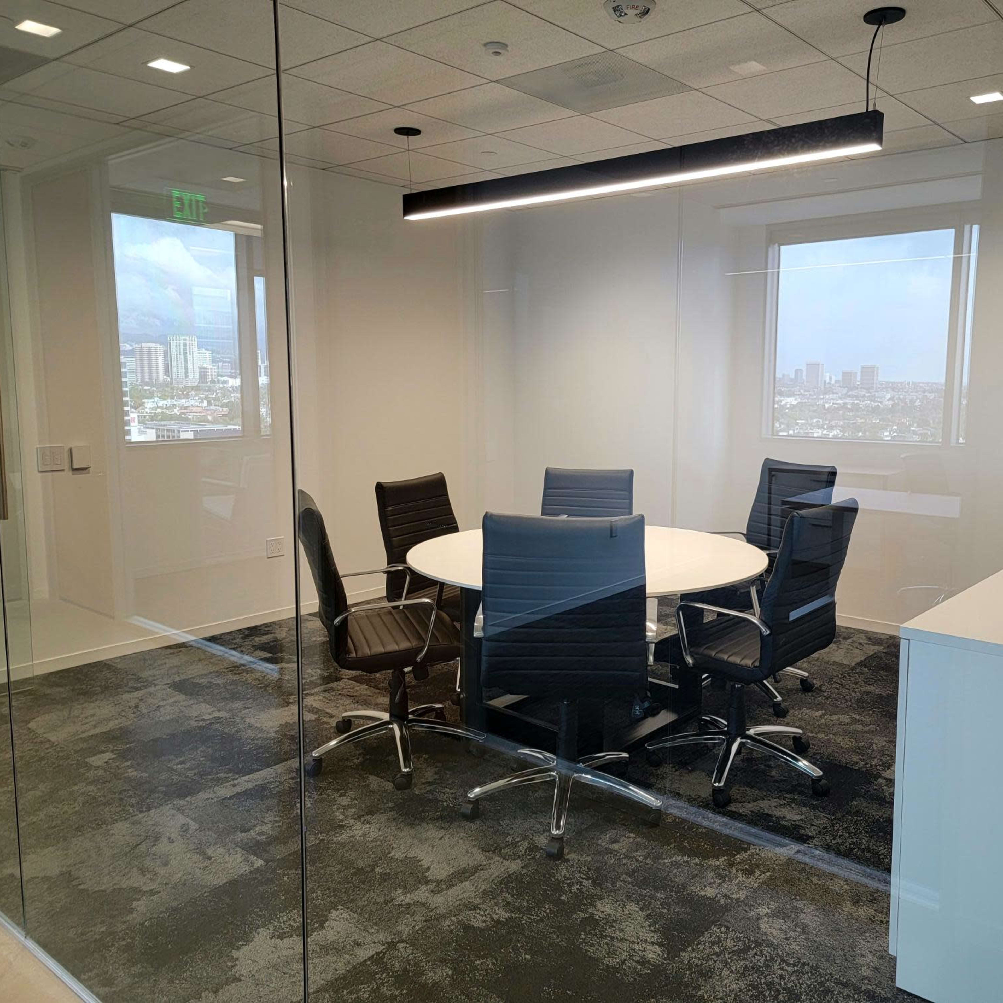 A modern glass-walled conference room with a round table surrounded by six black chairs and a minimalist side cabinet.