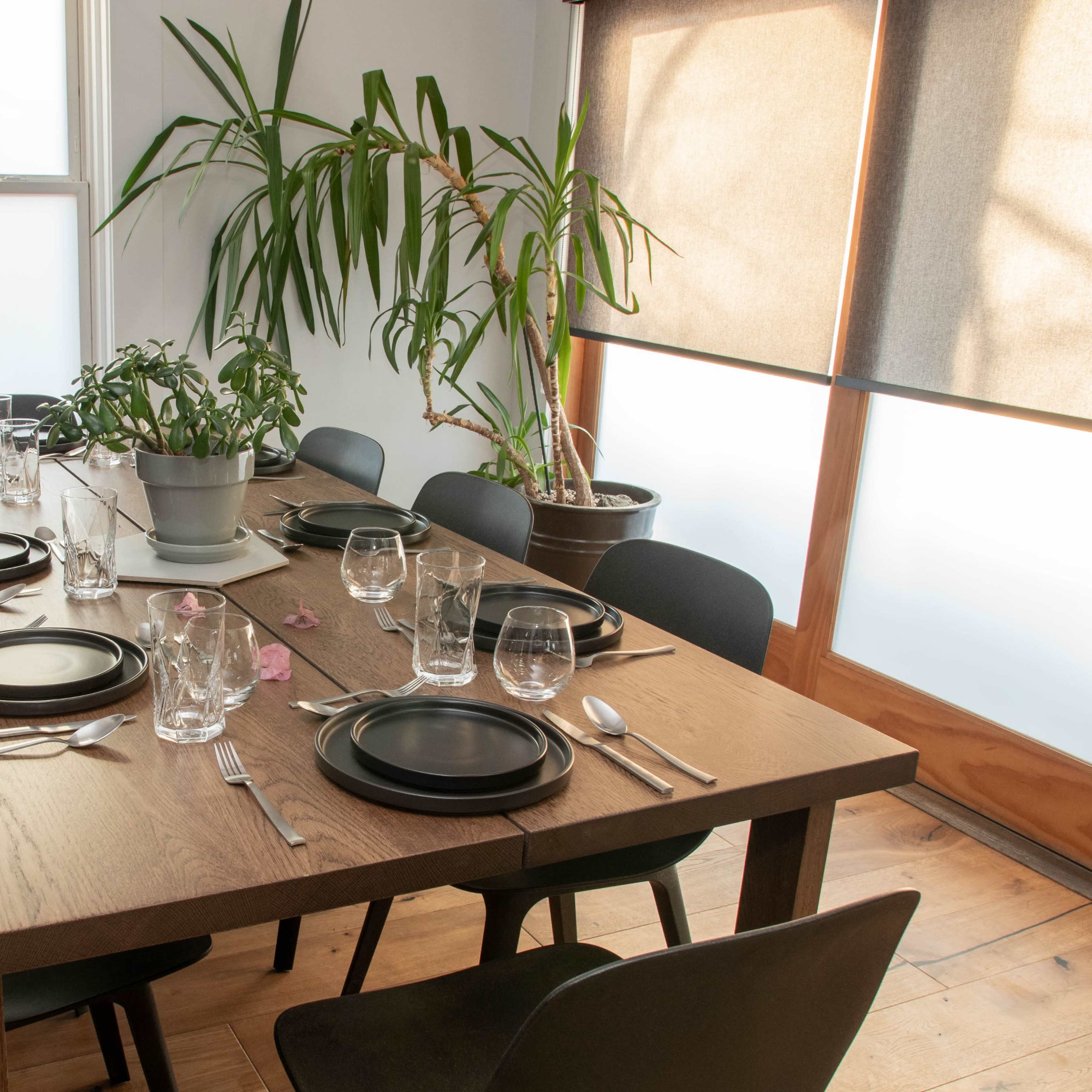A long wooden dining table is set with black plates, glassware, and silverware, surrounded by black chairs and flanked by potted plants.