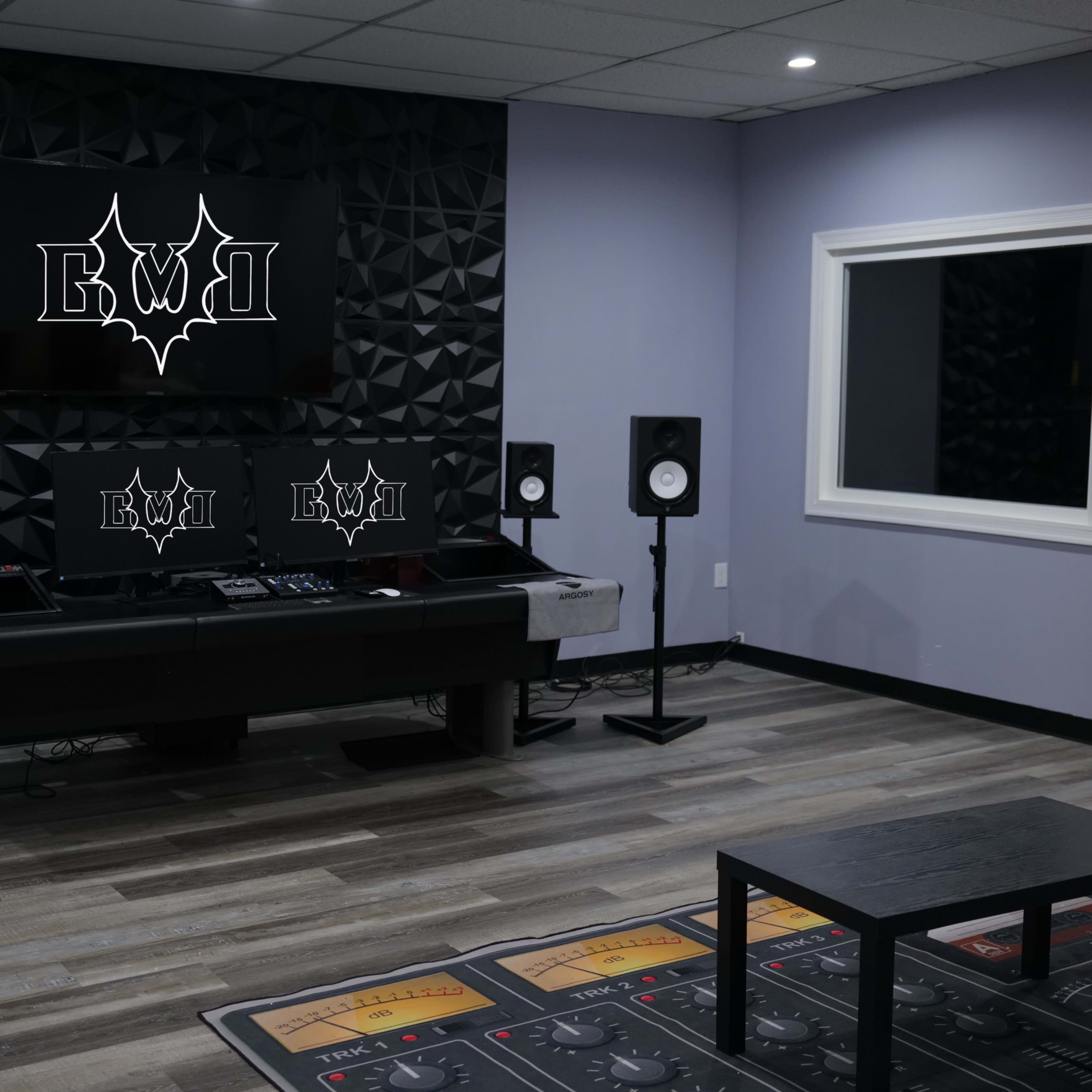 A modern music studio with dark acoustic paneling, a central mixing console, speakers on either side, and a large monitor displaying a logo on the wall.