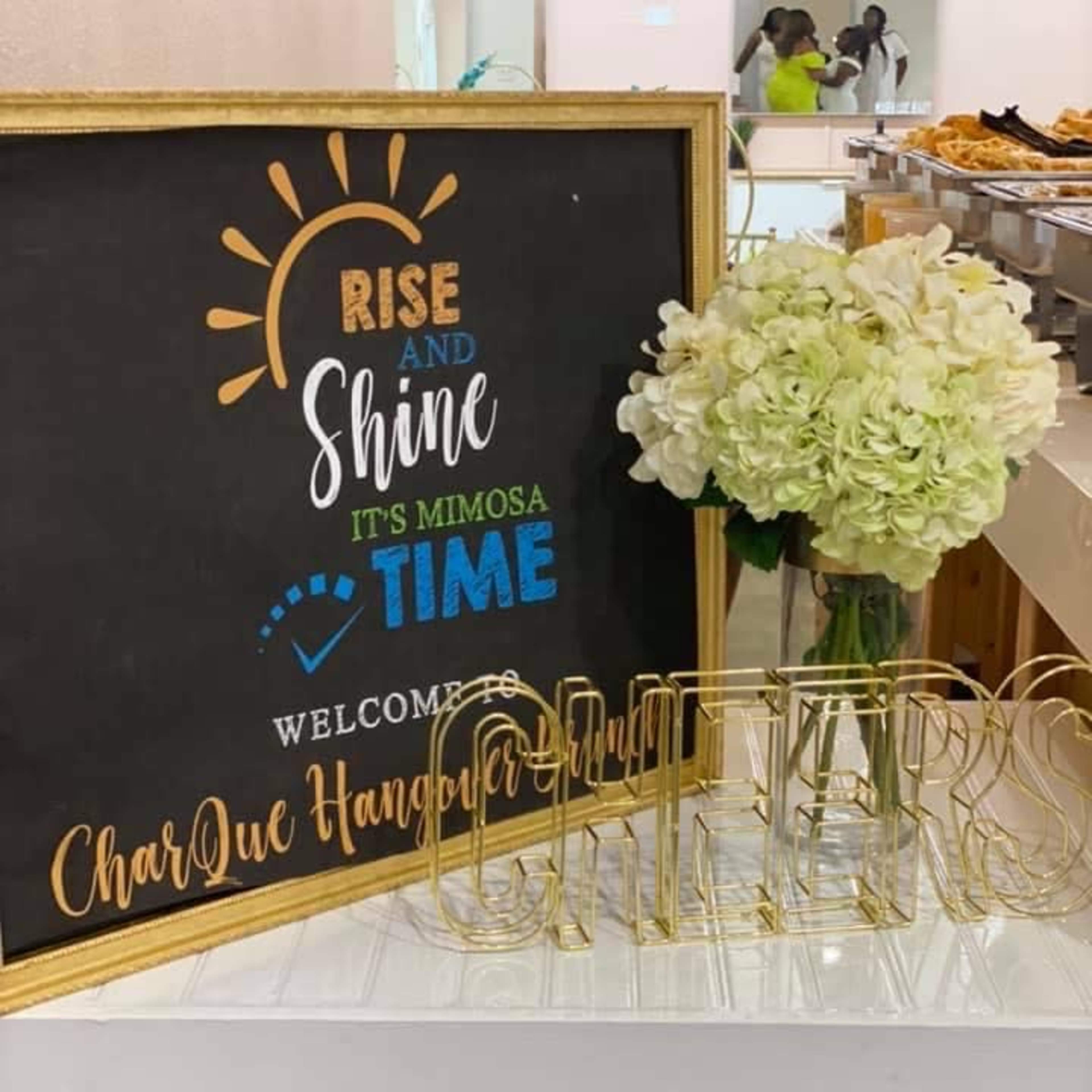 A framed sign reading "Rise and Shine, It's Mimosa Time" welcomes guests to an event, accompanied by a bouquet of white flowers and gold lettered decor.
