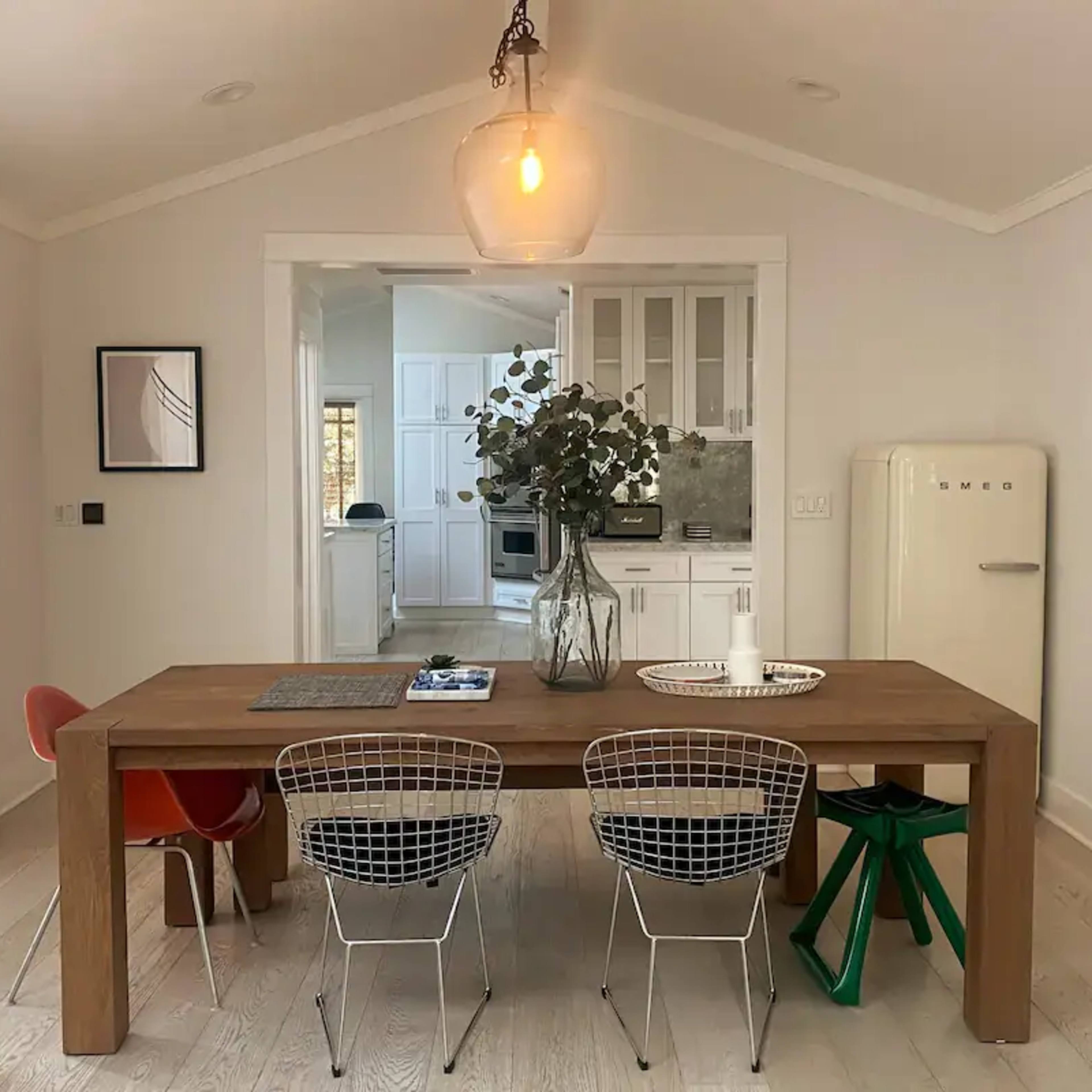 A wooden dining table is set with a vase of greenery and surrounded by colorful chairs in a light-filled room with a kitchen visible in the background.
