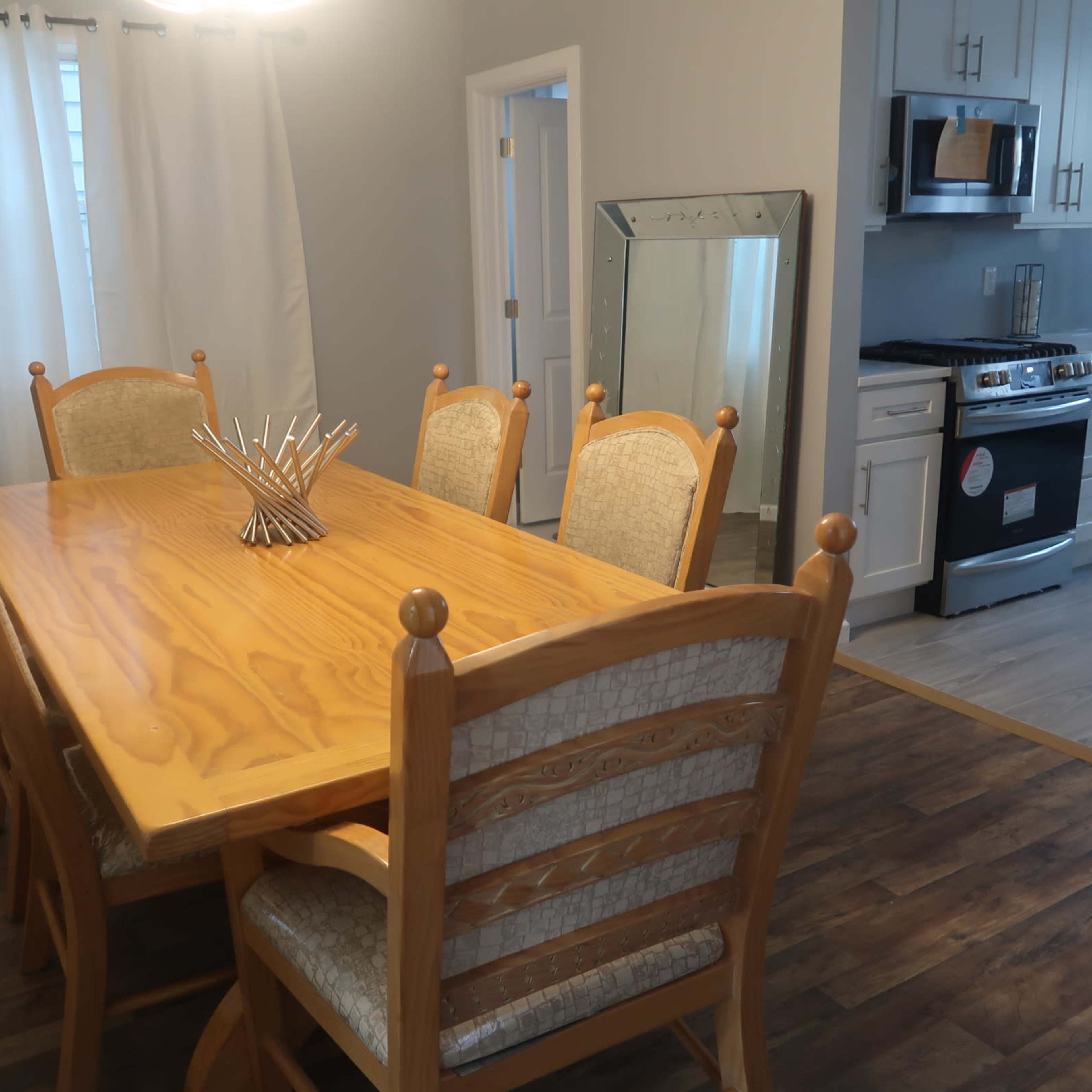 A wooden dining table with eight chairs is positioned beside a kitchen featuring stainless steel appliances and a mirror against the wall.