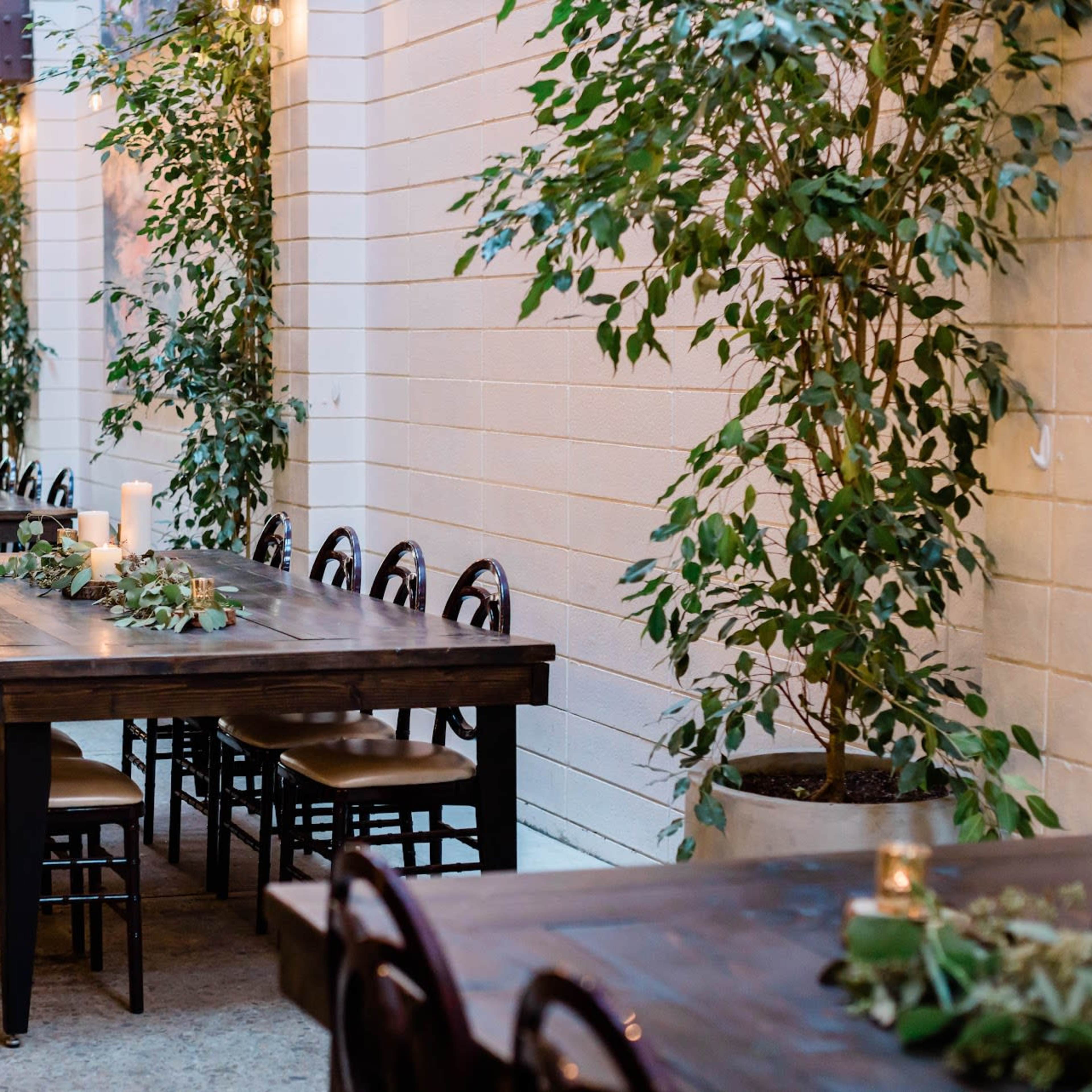 An outdoor dining area with long wooden tables, surrounded by potted plants and string lights overhead.