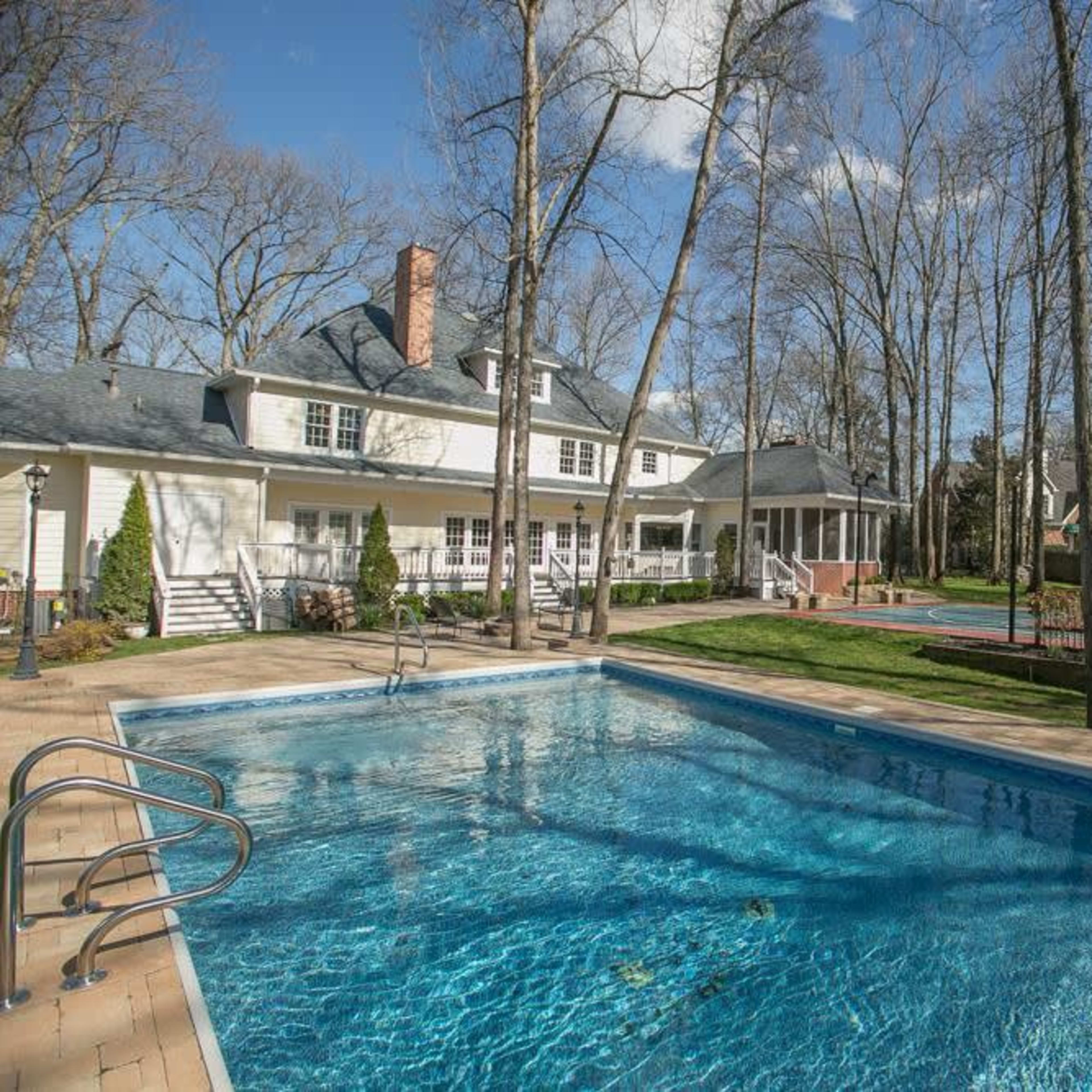 A backyard with a swimming pool surrounded by trees, a house in the background, and a tennis court to the side.