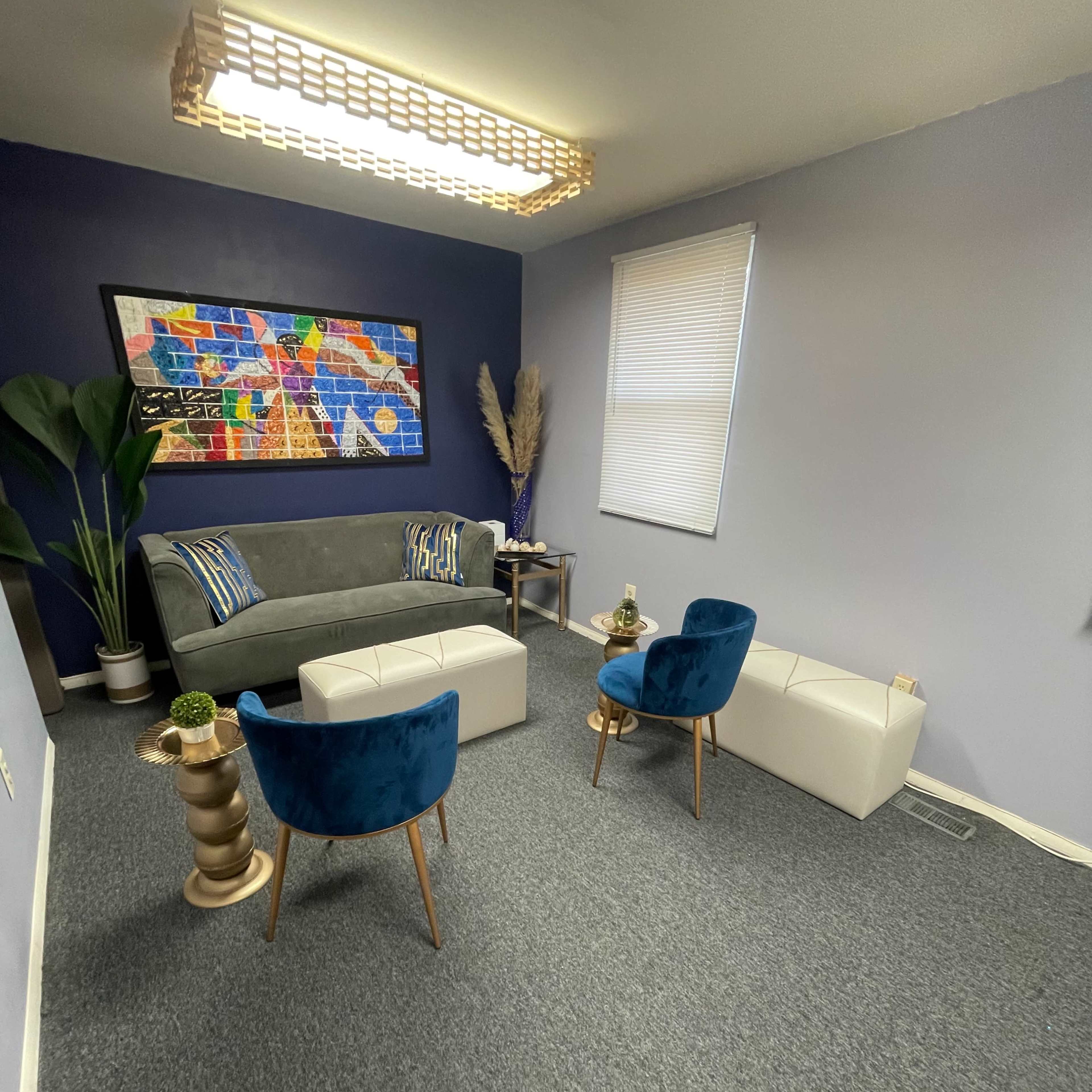 A small room features a gray sofa, two blue velvet chairs, a white coffee table, and colorful wall art illuminated by a modern light fixture.