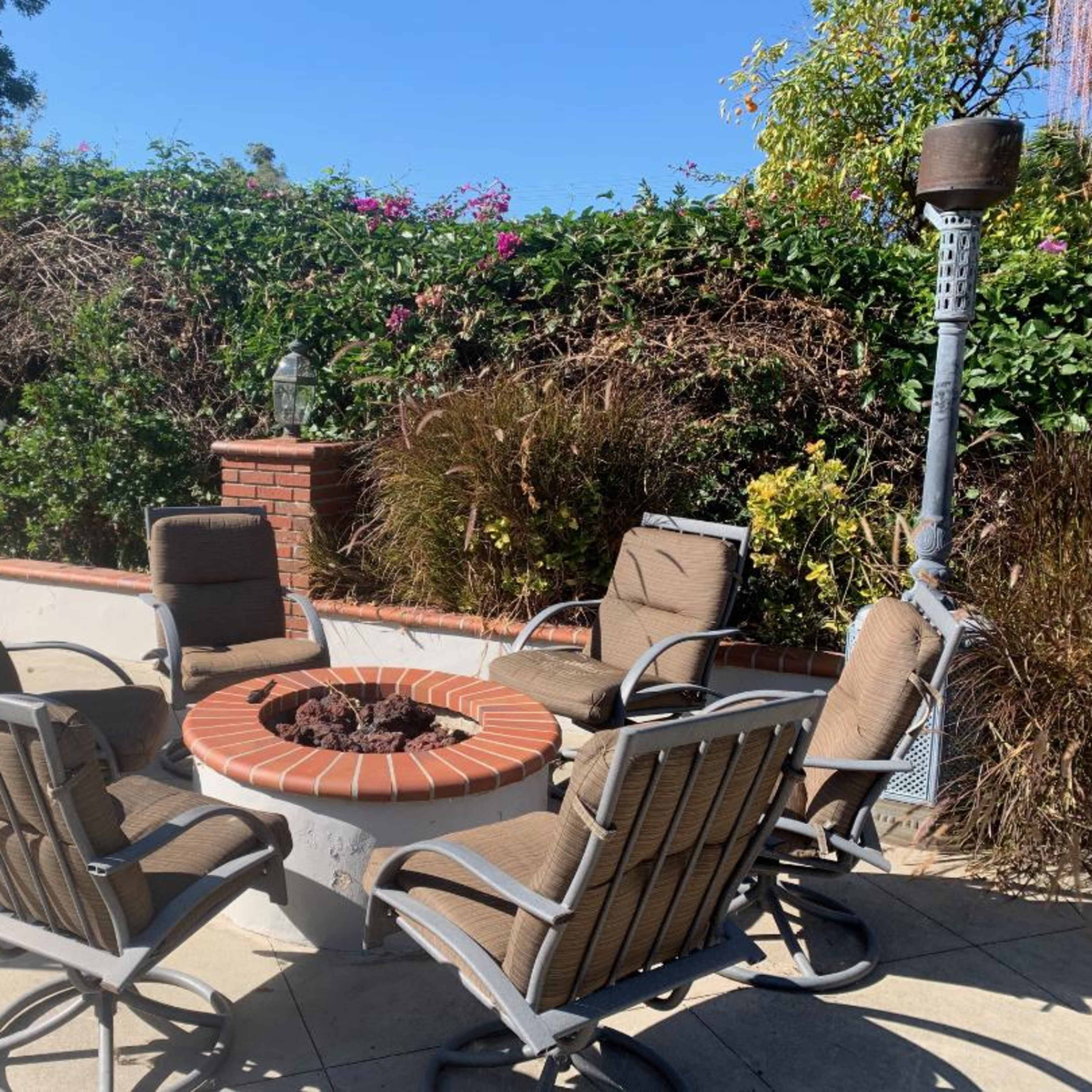 A circular fire pit surrounded by four chairs is set in a landscaped patio area with lush greenery in the background.