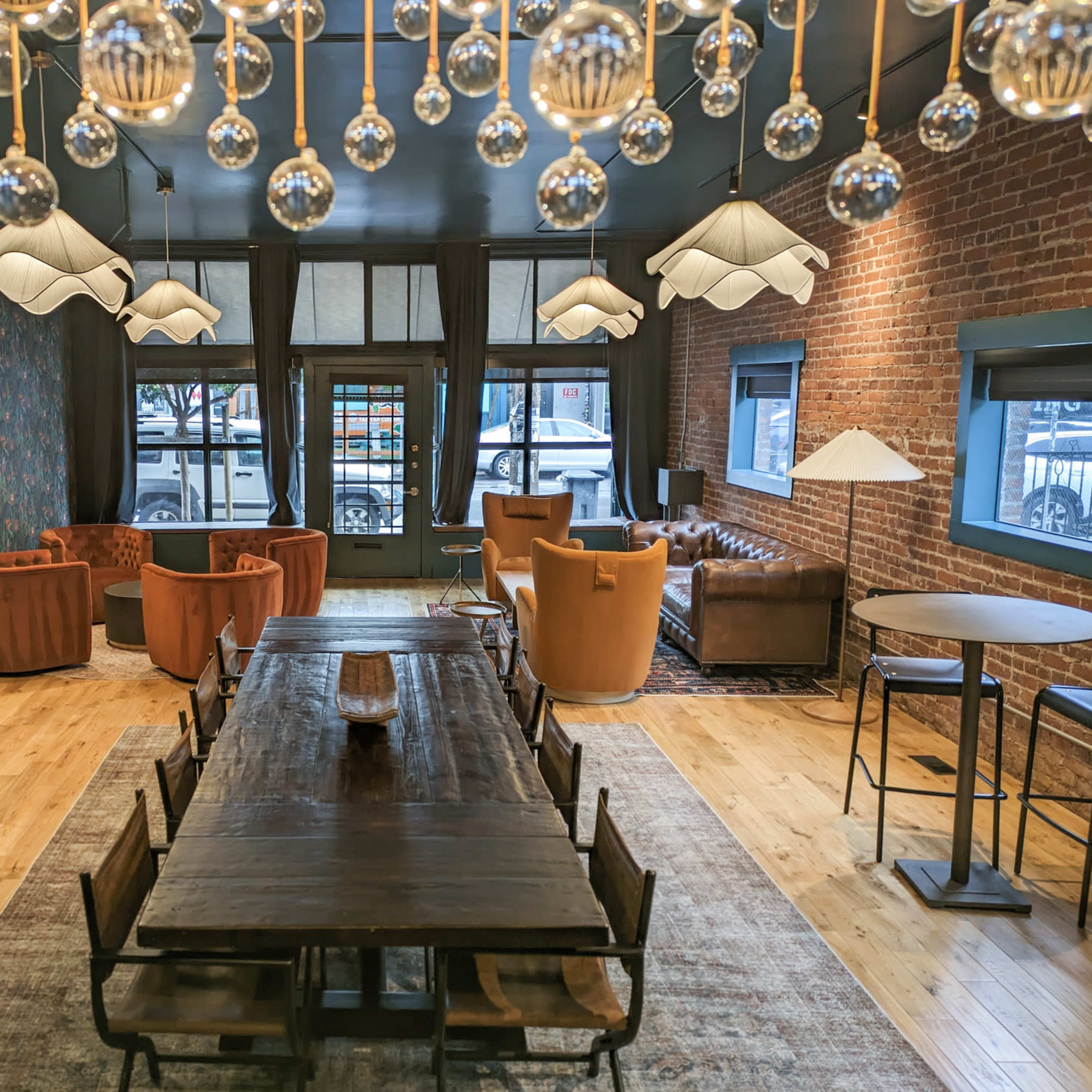 A spacious, modern lounge area featuring a long wooden table, several seating arrangements with armchairs, and unique light fixtures hanging from the ceiling.