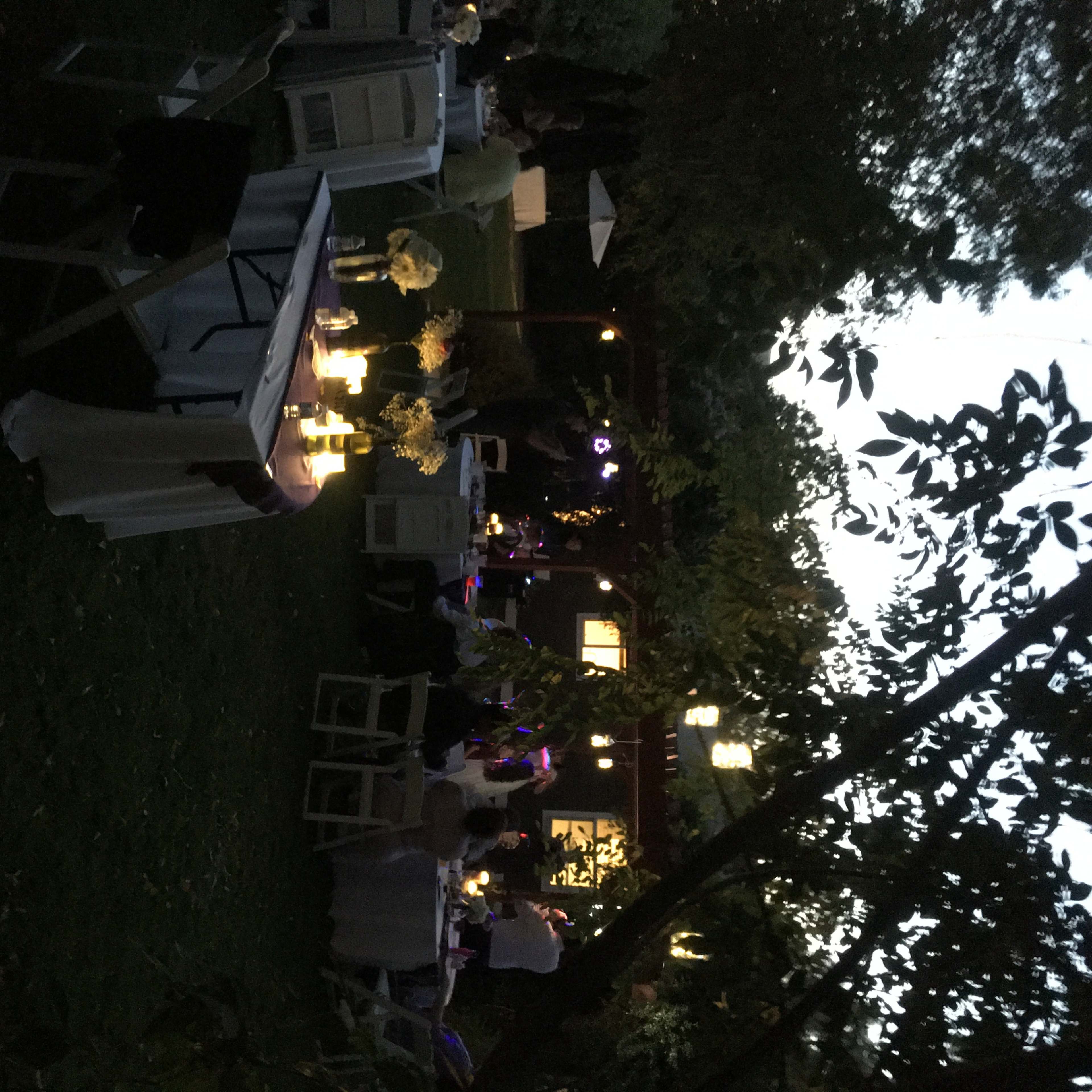 A garden setting with tables and chairs arranged for a gathering, illuminated by soft lights in the evening.