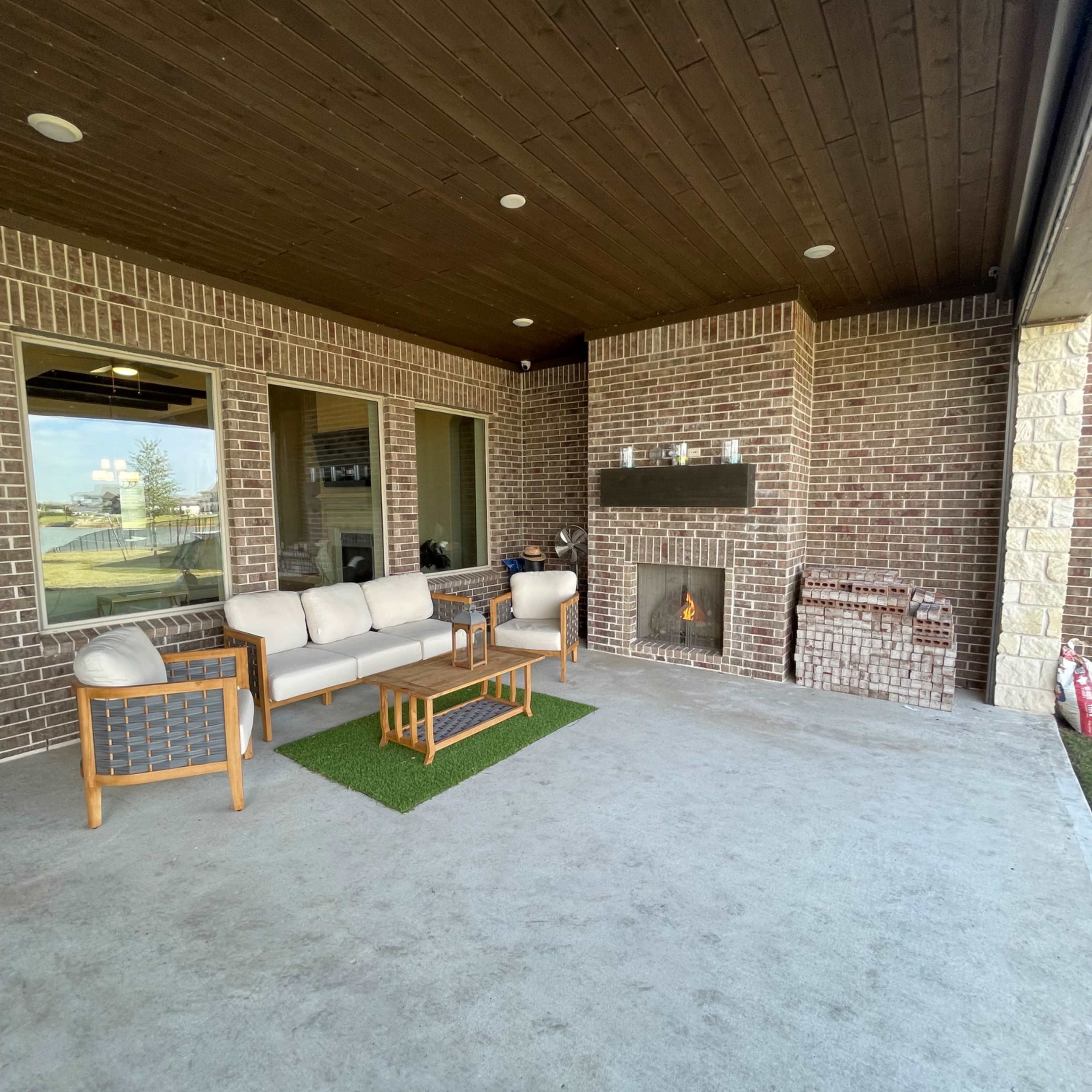 A covered patio area featuring a seating arrangement with a sofa, two chairs, a coffee table, and a fireplace surrounded by brick walls.