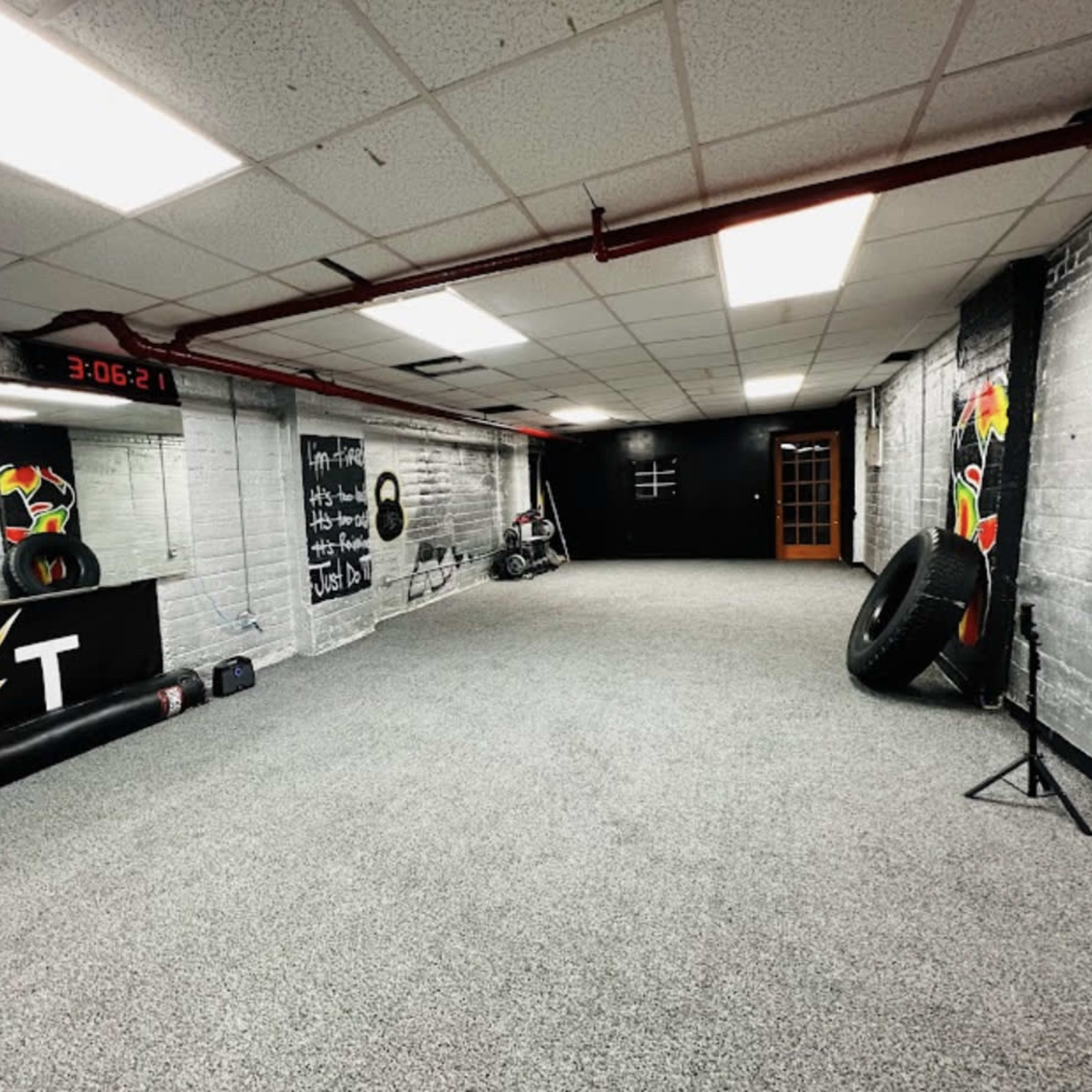 An empty gym or training room with gray carpet, white brick walls, and fitness equipment positioned along the sides.