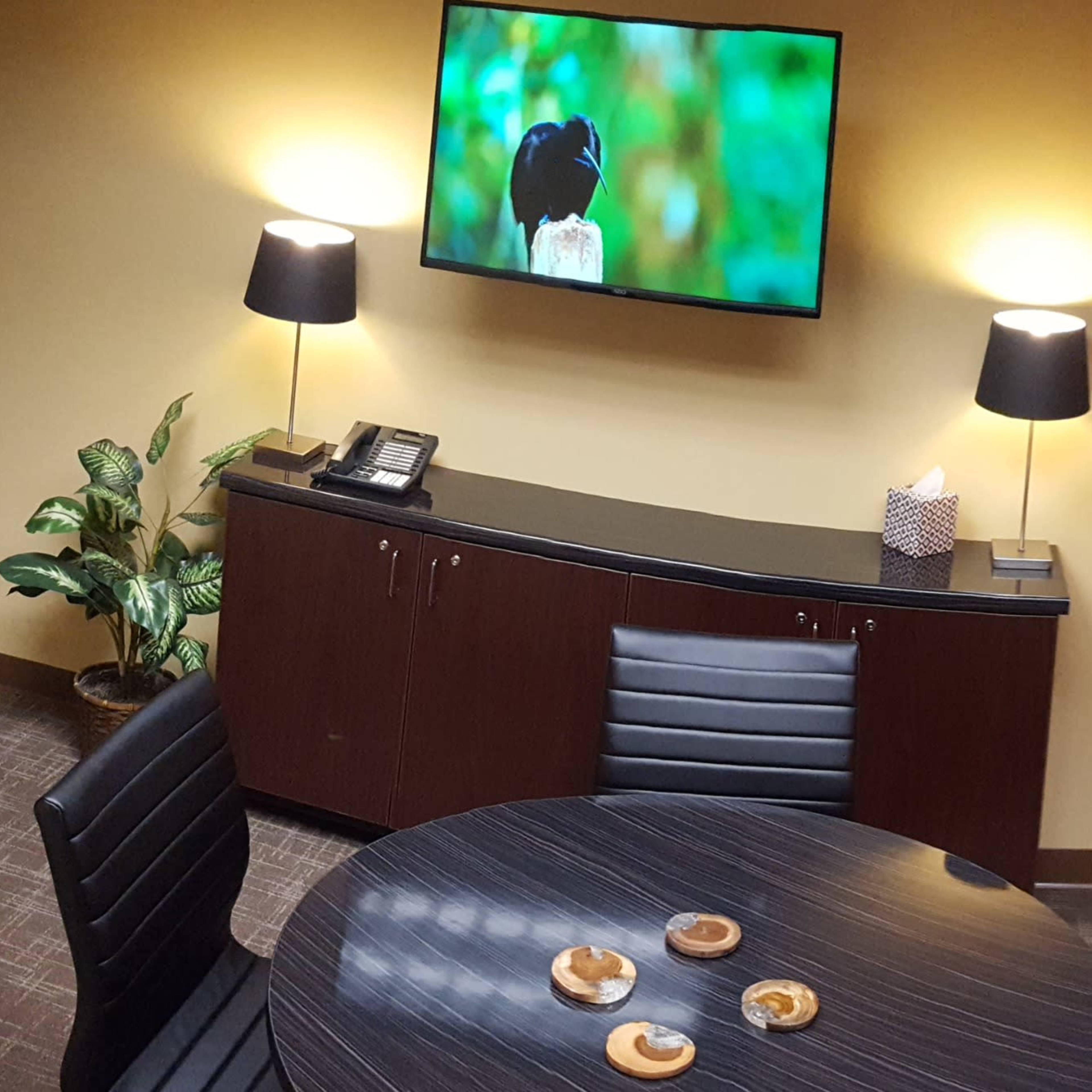 A small office meeting room features a round table with snacks, a television mounted on the wall, and decorations in warm colors.