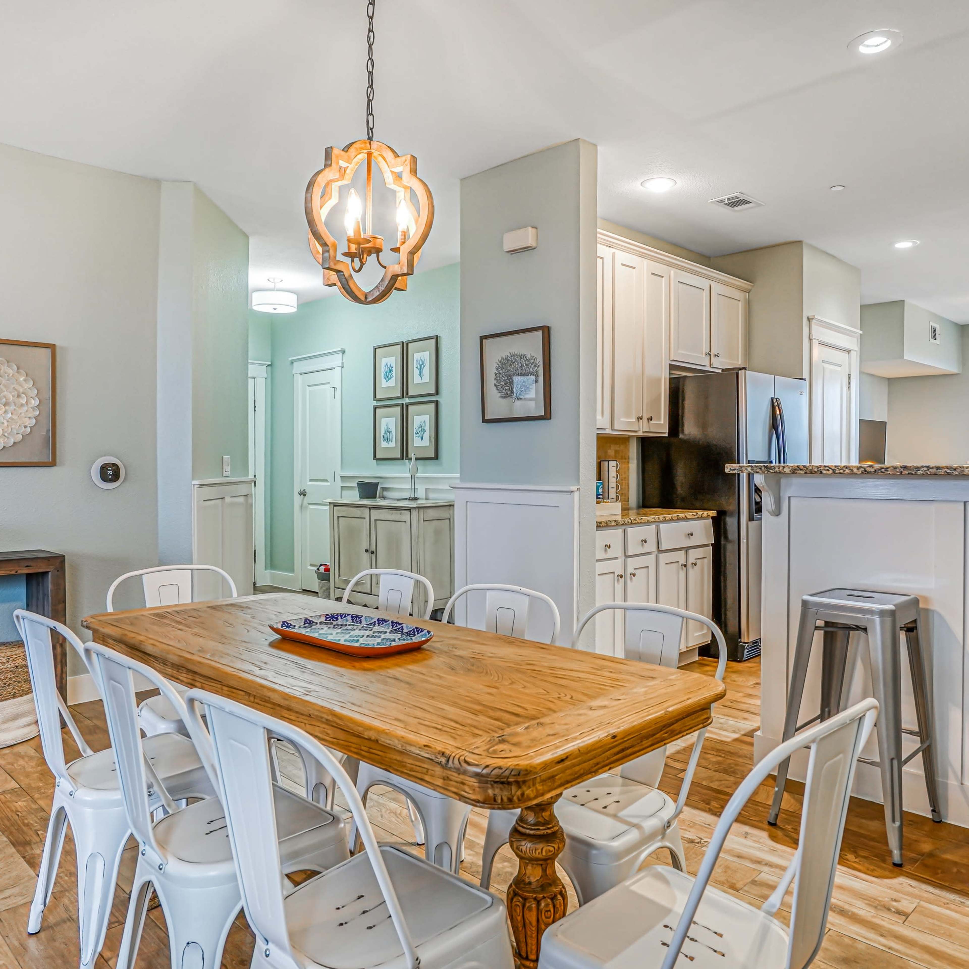 A dining area with a wooden table surrounded by white chairs, positioned near a kitchen with light-colored cabinets and a large window.