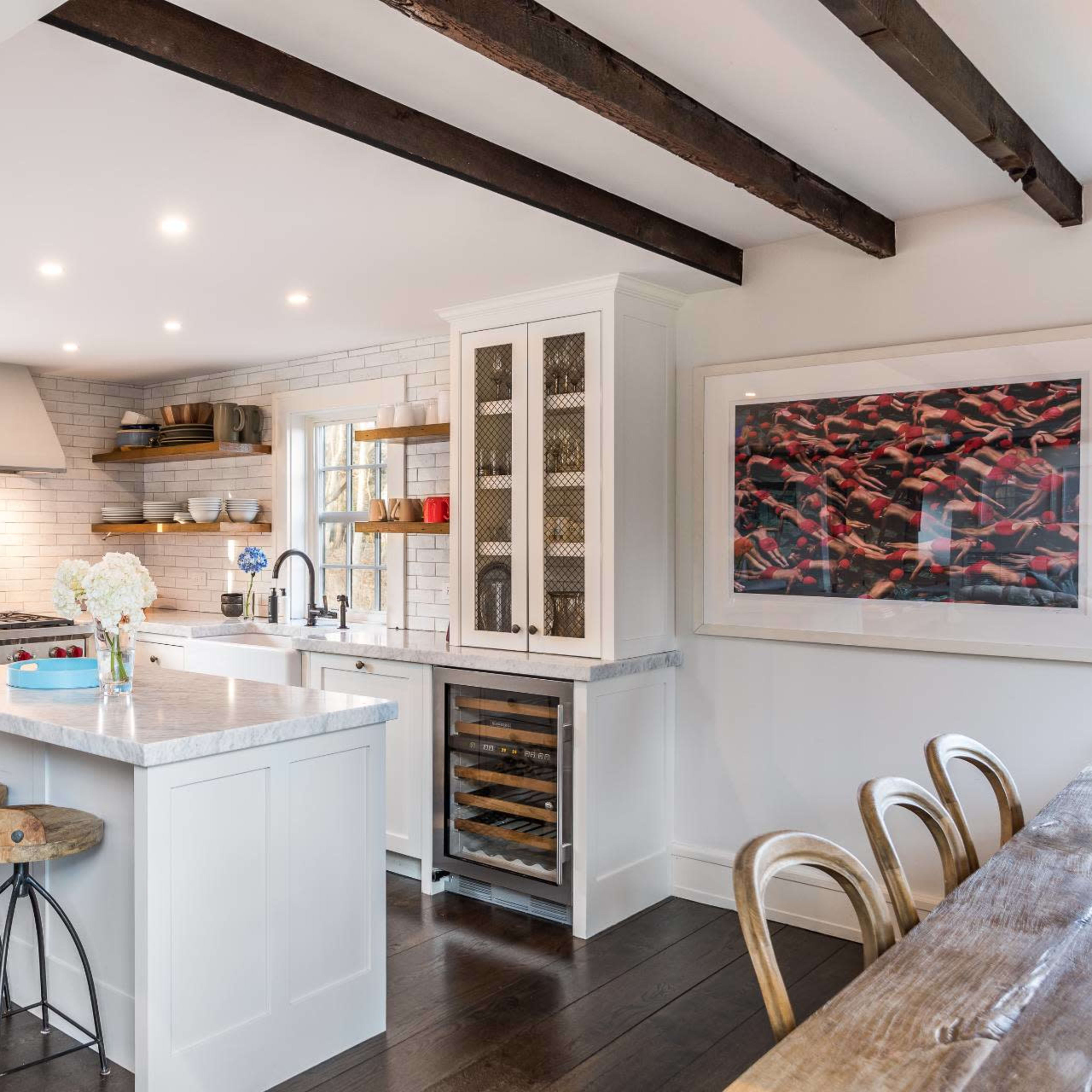 The kitchen features a central island with a marble countertop, wooden bar stools, and a combination of white cabinetry and exposed wooden beams.