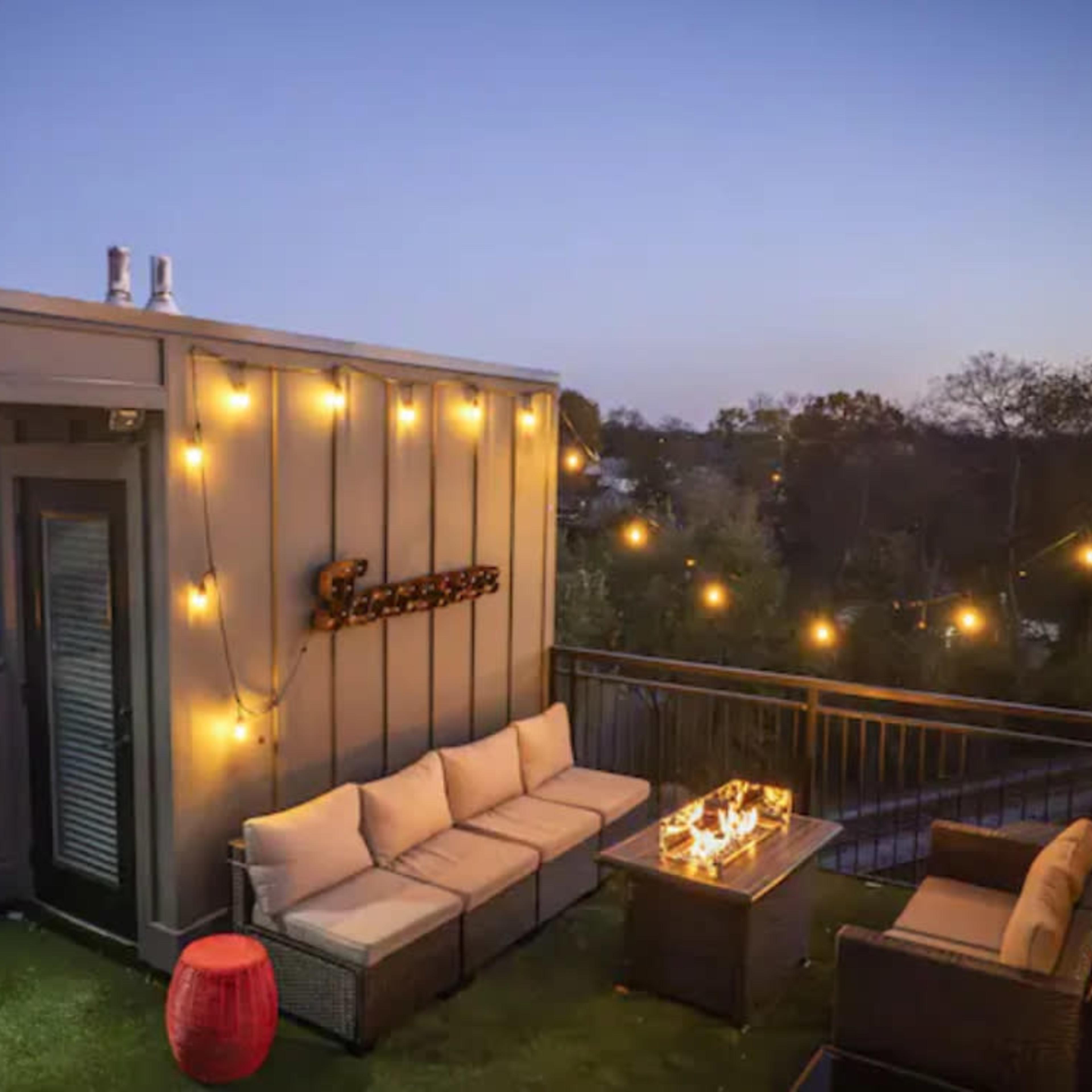 A cozy rooftop terrace features cushioned seating, a fire pit table, and string lights against a twilight sky.