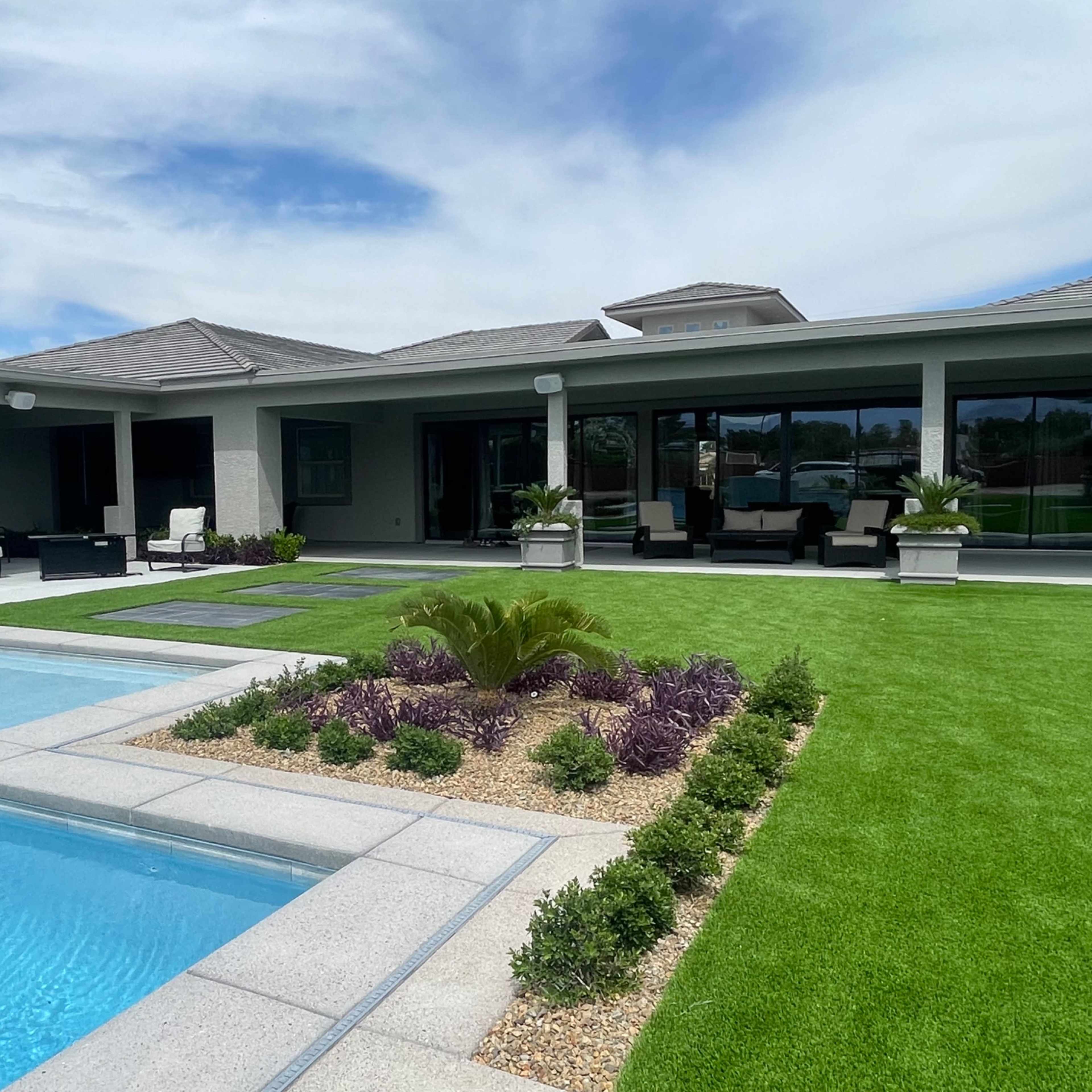 A modern home with a swimming pool and landscaped garden featuring green grass and various plants.